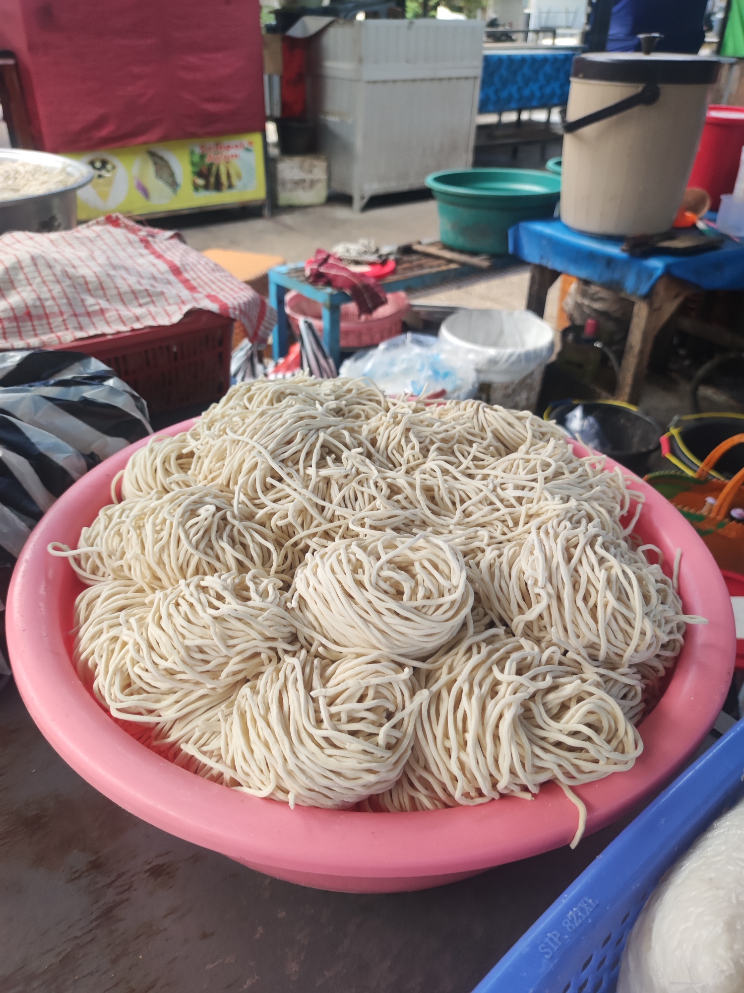 The image shows a pink plastic bowl filled with bundles of fresh, uncooked noodles. The bowl is resting on a surface with a textured appearance, possibly a table or countertop. In the background, there is an outdoor food preparation area. Visible elements include a food stall with a red awning, various containers (buckets, basins), and a blue table. Some people are partially visible in the background. The photo was taken in Gianyar, Indonesia.