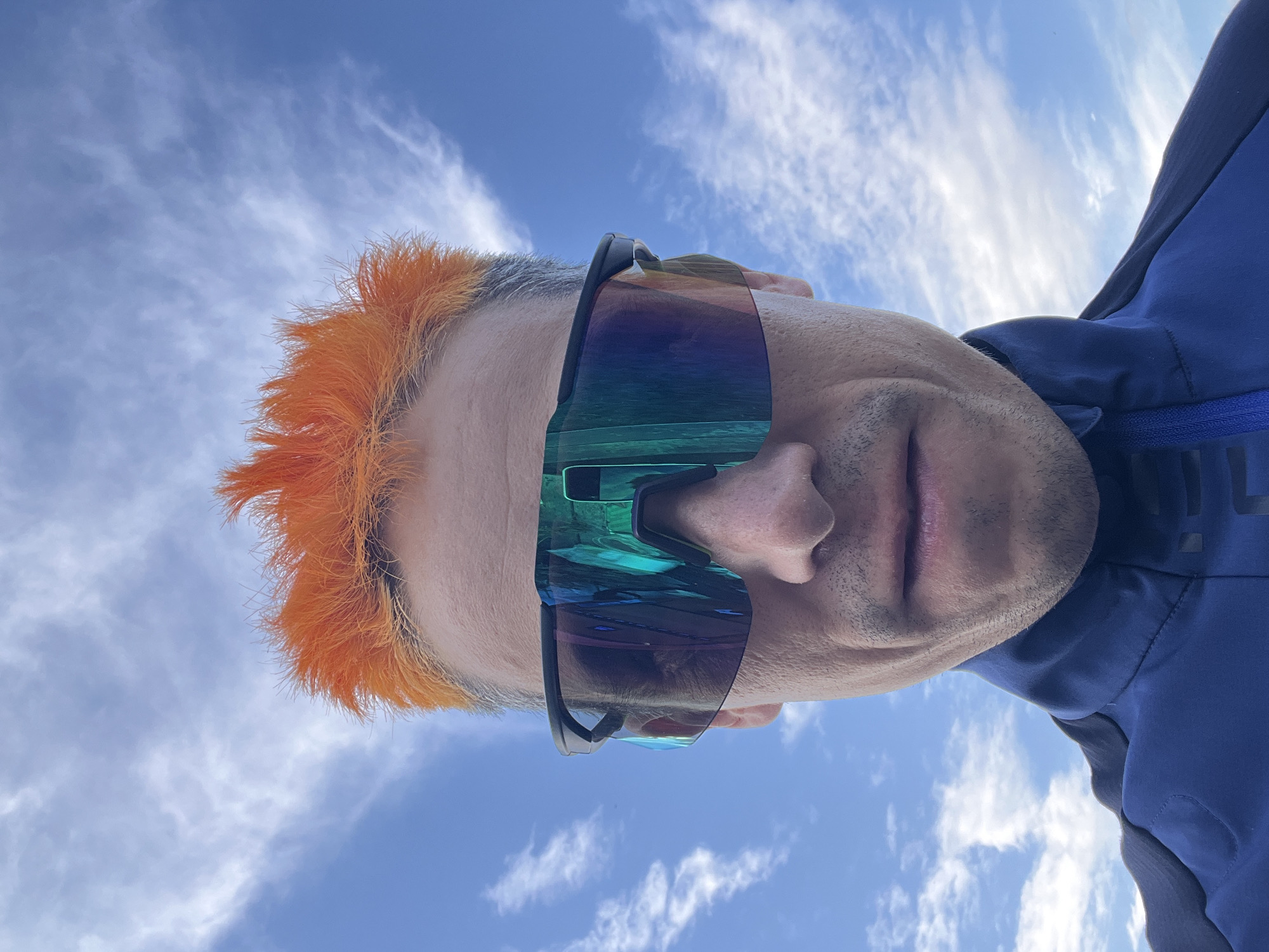 A close-up, low-angle, eye-level outdoor selfie shows a person's face and upper torso against a bright blue sky with scattered white clouds. The subject has vibrant, spiky orange hair and is wearing large, dark-framed sporty sunglasses with iridescent, multi-colored lenses that reflect the sky. The person's face appears weathered, with a slight mustache and stubble. They are wearing a dark blue jacket with a raised collar. The background is a clear sky, typical of an outdoor setting. No specific location in Chaikovsky, Russia can be identified from the visual information provided.
