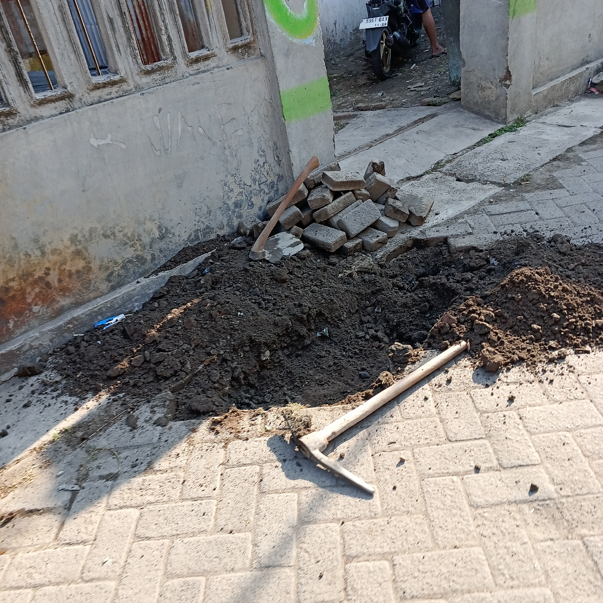 The image depicts a scene in the city of Wedoro, Indonesia, showing evidence of construction or repair work. A pile of dark soil is central to the image, adjacent to a section of cobblestone paving that has been disturbed. Two tools are visible: a shovel or hoe with a wooden handle and a metal head lying on the cobblestones, and a shovel with a wooden handle and a metal pointed end resting on the soil pile. A stack of grey paving stones is also present near the soil. In the background, a wall of a building, possibly under construction or renovation, shows signs of wear and has some graffiti or paint marks, including a bright green curved line. A motorcycle with a license plate reading "P 5961 DAX" is parked near the building, and a person's legs and feet are visible near it. The environment appears to be an outdoor urban or suburban area.