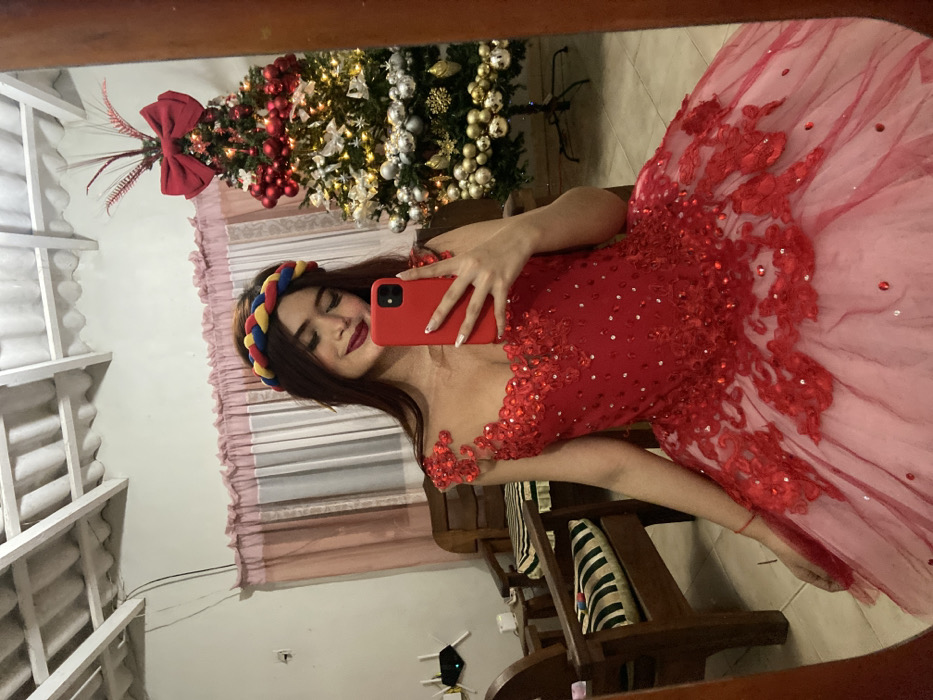 A young woman in a red, elaborately decorated dress poses for a selfie in a mirror. She wears a colorful braided headband. A Christmas tree stands in the background with red and gold ornaments. The room also contains wooden furniture, including chairs with striped cushions. The mirror reflects part of the ceiling, which appears to be corrugated.