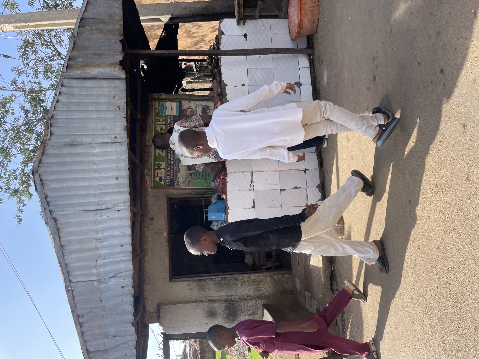 In Barakin Akawo, Nigeria, two young boys walk along a dusty path in front of a corrugated metal-roofed market stall. The boy on the left wears a dark purple tunic and trousers, with light green slide sandals. The boy on the right is dressed in a white long-sleeved shirt and light-colored trousers, with dark slide sandals. Behind them, a man in a patterned shirt stands within the market, which displays white tiled walls and various goods, including what appears to be meat. A blue scale is visible inside the stall. The ground is a mix of packed earth and asphalt. Overhead, a bright sky is partially obscured by tree branches. An orange plastic basin sits near the right edge of the frame.