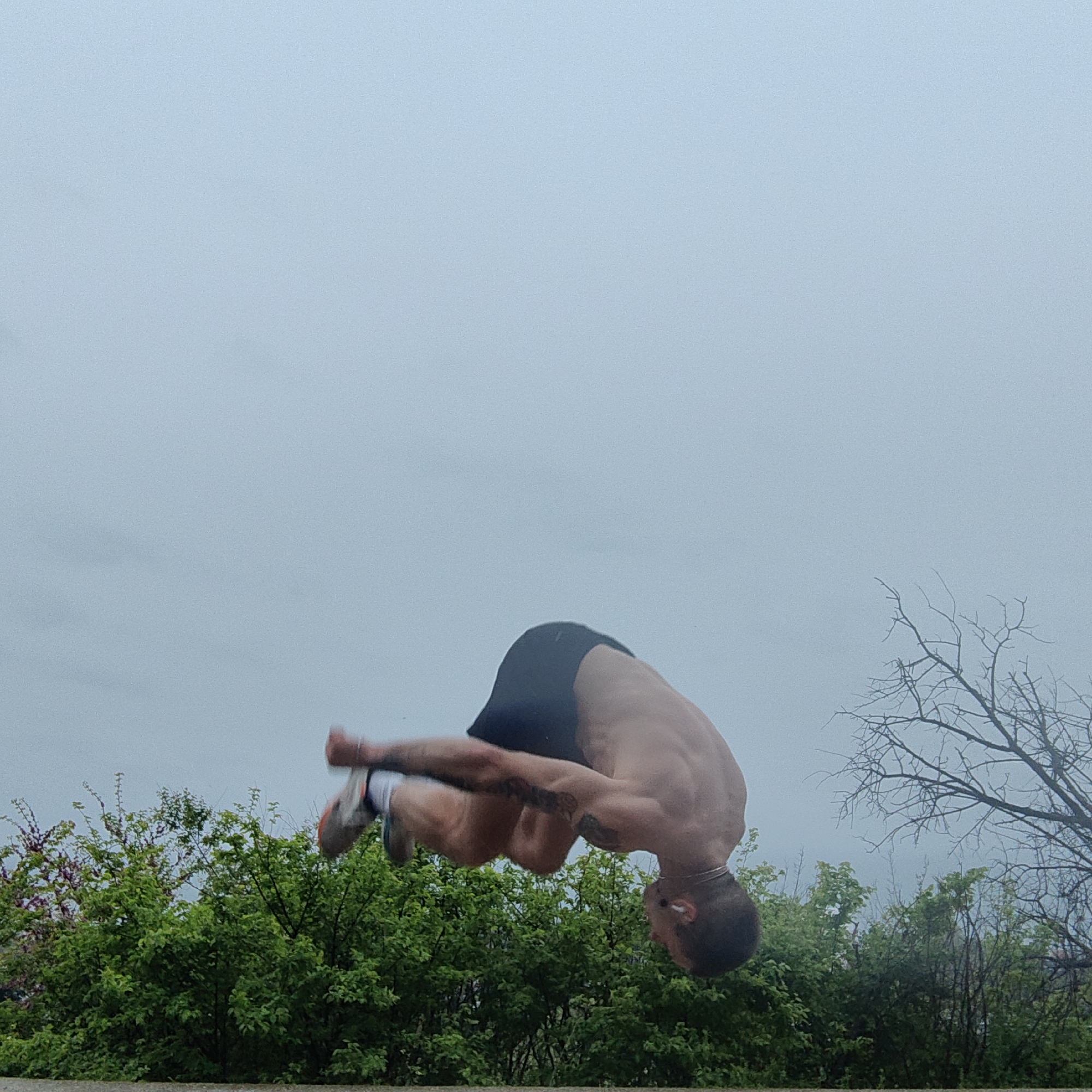 An outdoor, mid-action photograph captures a muscular man performing a backflip against an overcast sky in Varna, Bulgaria. The man is shirtless, wearing dark shorts, light-colored athletic shoes with white socks, and appears to have a silver chain around his neck and possibly earbuds. His body is tightly tucked, with his head facing downwards, indicating he is in the process of rotating through the flip. Visible tattoos adorn his left arm.
The upper two-thirds of the image are dominated by a soft, light blue-grey overcast sky, suggesting cloudy daytime weather. The lower third of the frame features a lush expanse of green bushes and trees, with a prominent bare-branched tree on the right side, hinting at a natural outdoor setting like a park or open field. A narrow, dark strip at the very bottom suggests a ground or wall edge. The image conveys athleticism and dynamic movement within a serene, natural environment. There is no visible text.