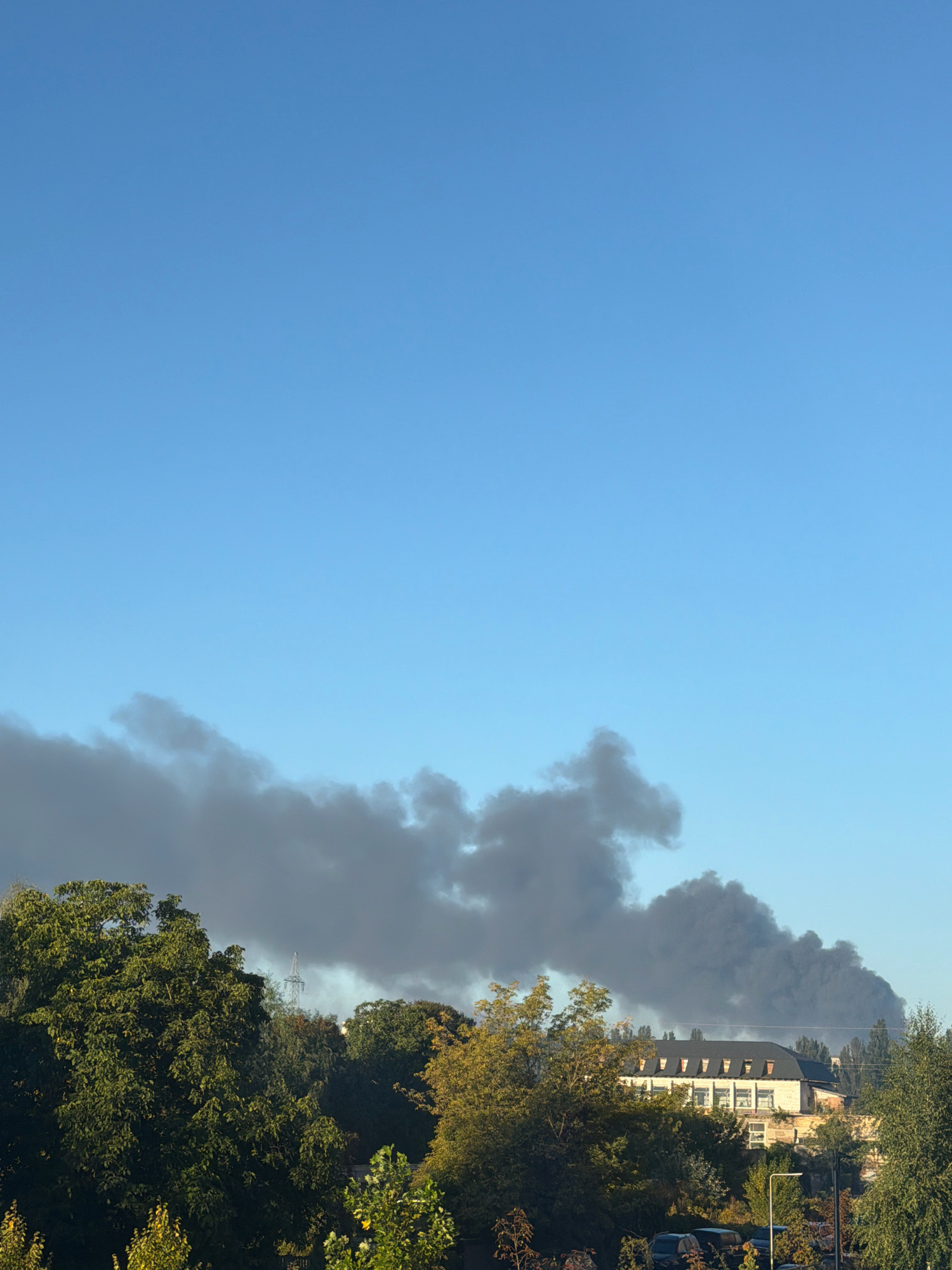 In Kyiv, Ukraine, a large plume of thick, dark smoke billows into the clear blue sky, indicating a significant fire or event. The smoke originates from beyond a line of lush green trees and a building with a dark roof and multiple dormer windows. The building appears to be a multi-story structure, possibly an industrial or institutional building, with numerous windows visible. The trees in the foreground are dense and verdant, suggesting a temperate climate. The overall scene depicts a clear day disrupted by the dramatic visual of smoke rising from an unseen source in the distance.