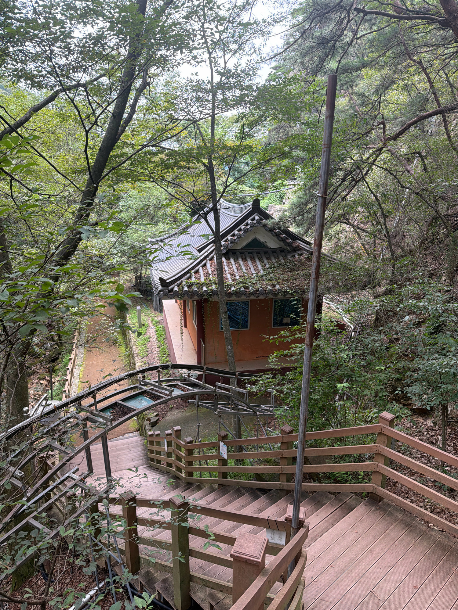 A high-angle, medium shot captures a wooden staircase and an old, traditional Korean-style house nestled in a lush, green forest. The staircase, made of weathered brown wood, features sturdy railings and descends towards the left of the frame, with a metallic structure arching over a portion of it. The house, with its distinctive tiled roof and orange walls, is situated further into the woods, partially obscured by trees and foliage. A narrow, dirt path runs alongside the house and leads into the dense forest. The overall scene is one of a tranquil, natural setting, possibly a tourist destination or a secluded retreat within Dongducheon-si, South Korea.