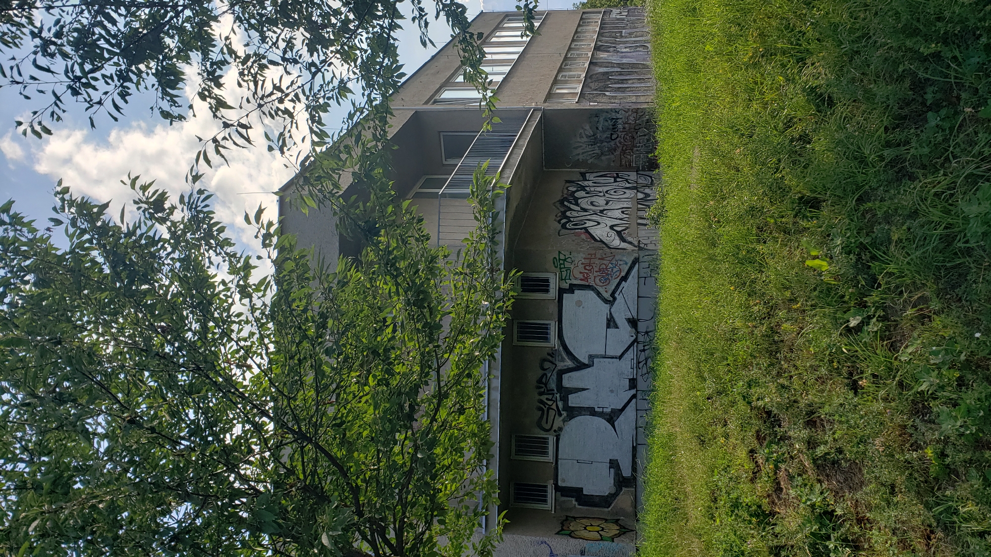 The image shows a two-story building with a balcony and several windows. The lower part of the building is covered in graffiti, including large, stylized lettering and more complex designs. The building is situated on a grassy slope, and trees with green leaves partially obscure the view. The sky above is partly cloudy. The building is located in Prešov, Slovakia.