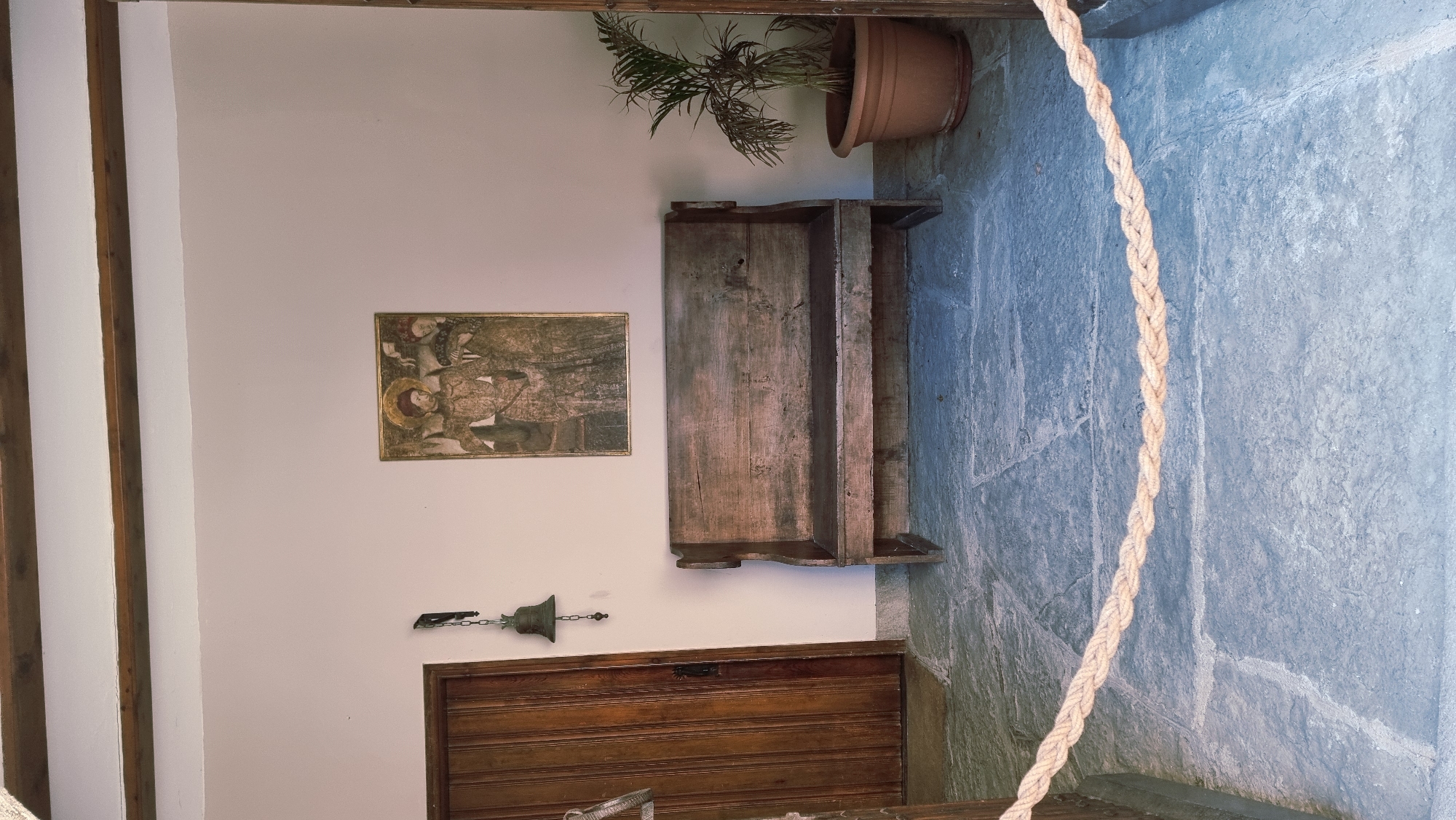 This is a full, eye-level, vertical shot of an entryway or courtyard in Rupit i Pruit, Spain. The scene features a rustic wooden bench with a weathered finish positioned centrally against a pale pink wall. Above the bench hangs a framed painting depicting two figures in historical attire. To the left of the bench is a tall, wooden door with vertical planks. A small, dark-colored bell hangs by a chain from a bracket on the wall to the left of the door. To the right of the bench, a green plant in a terracotta pot sits on the stone floor. The floor is made of large, irregularly shaped stone slabs, giving the impression of an old or natural setting. A thick, braided rope barrier runs horizontally across the lower portion of the image, separating the viewer from the depicted scene. Wooden beams are visible along the top of the frame, suggesting an overhead structure like a porch or covered walkway. The lighting is bright and natural, indicating daytime.