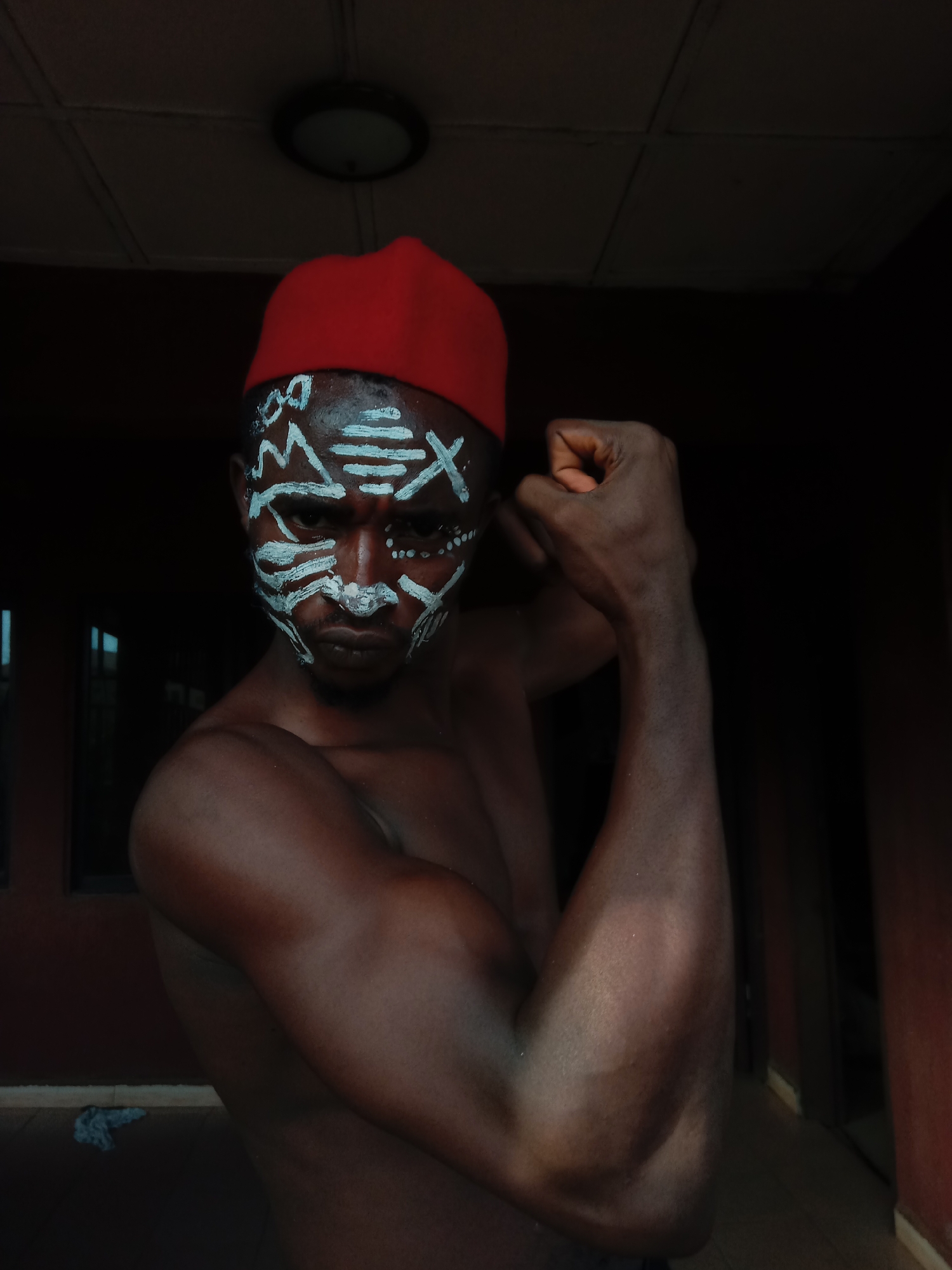 A shirtless dark-skinned man with white tribal face paint and a red fez stands indoors in Nkpor, Nigeria. He is flexing his arms. A ceiling light is visible above him. A window and a red-painted wall are behind him. A piece of cloth lies on the floor behind him.