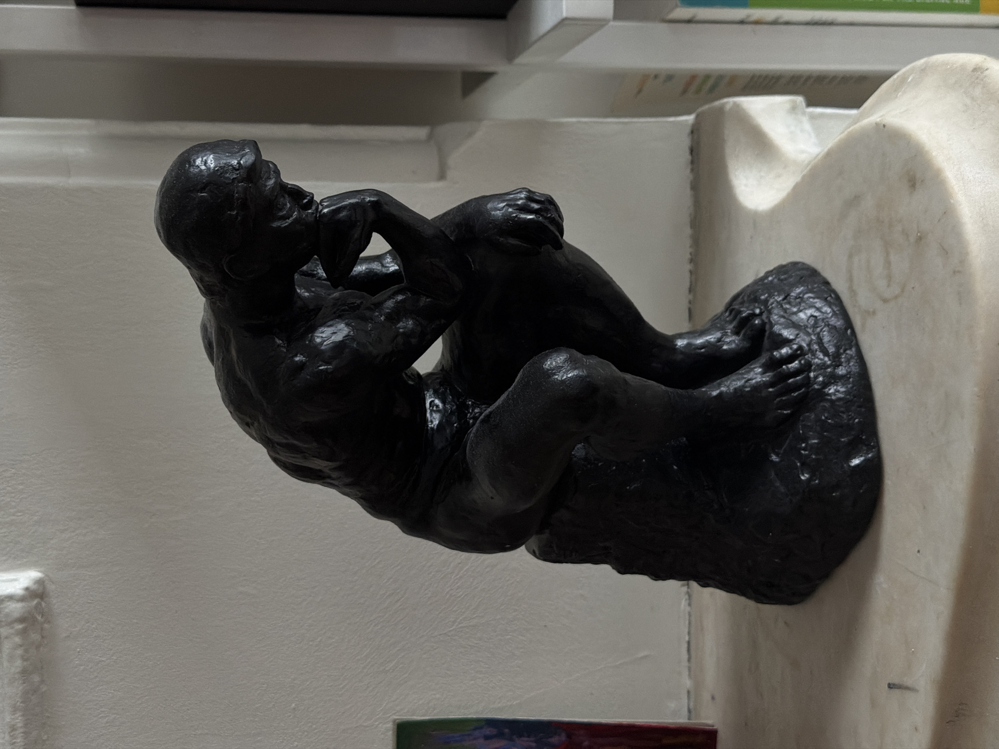 A bronze sculpture of "The Thinker" by Auguste Rodin sits in a museum setting. The dark bronze sculpture is mounted to a light-colored marble or stone base. The sculpture depicts a nude male figure in a seated position with his chin resting on his right hand. The man is in deep contemplation. The setting appears to be inside a museum, as suggested by the painted walls, architectural details, and what seems to be a bookshelf in the background. The location is the Iris and B. Gerald Cantor Foundation.