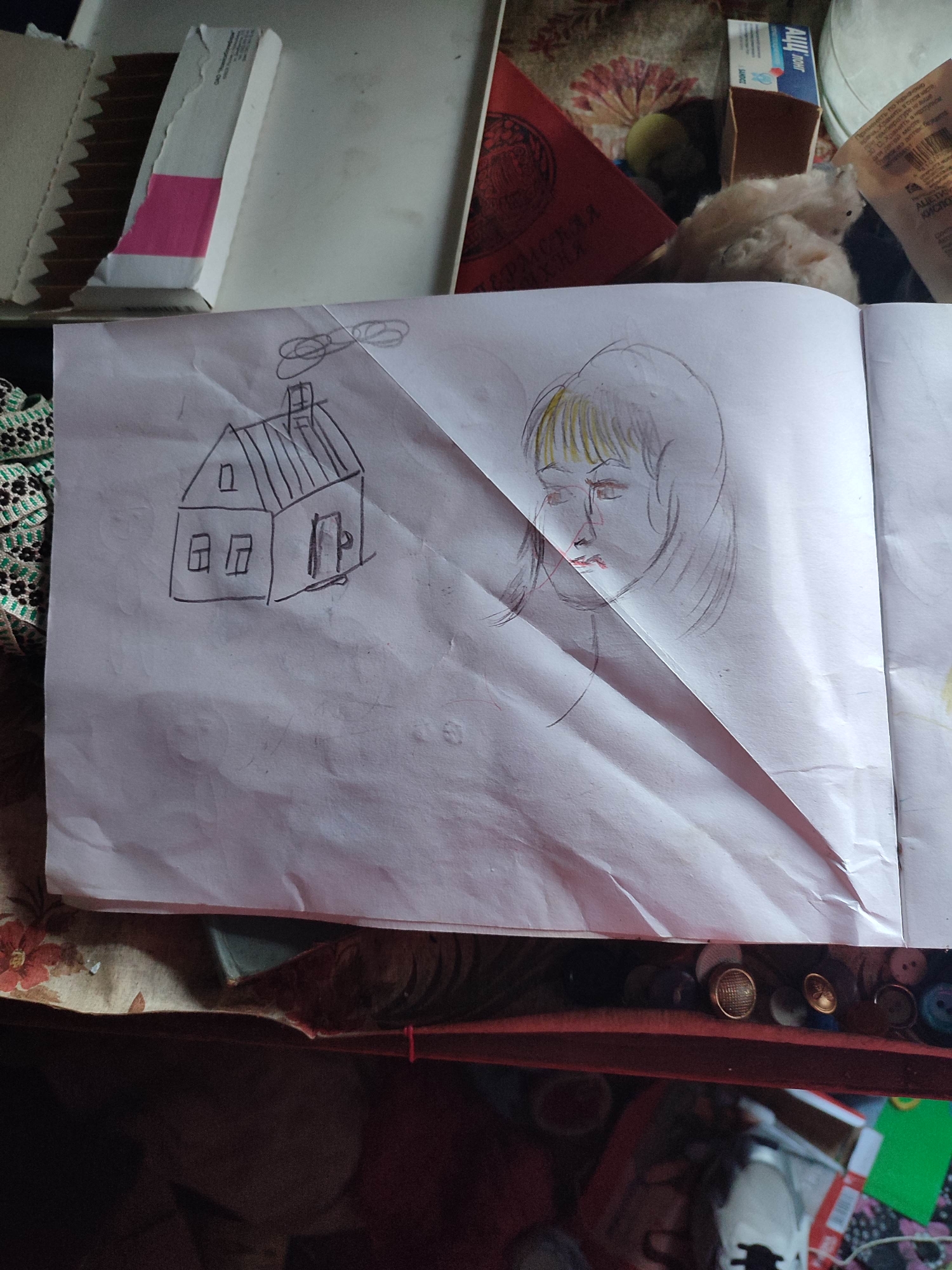 The image shows a spread of what appears to be an open notebook or sketchbook. On the left page, a child's drawing of a house with a chimney and a few circular shapes above it, possibly representing clouds or a helicopter, is visible. The house is drawn with simple lines, showing windows and a door. On the right page, a sketch of a person's face is drawn, with yellow and brown colored hair, and red on the lips and cheeks. The drawing is slightly abstract, with bold lines and a visible shadow cast across it.
The notebook is placed on a surface that appears to be a table or desk, with various items scattered around. To the top left, a cardboard box with a pink and white label is partially visible. There are also glimpses of fabric with a floral pattern, a red book with Cyrillic text, a small fluffy object, and a blue and white box with some text. The bottom of the image shows a collection of buttons and spools of thread, suggesting a crafting or sewing area.
The overall setting seems to be indoors, in a somewhat cluttered but lived-in space, possibly a home. The lighting suggests it is daytime, but no specific time of day can be determined. There are no clear indications of weather conditions or a specific outdoor location. The drawings themselves evoke a sense of childhood creativity. The visible text includes Cyrillic lettering on the red book and the blue and white box, with some labels like "SANDOZ" and "PERM".