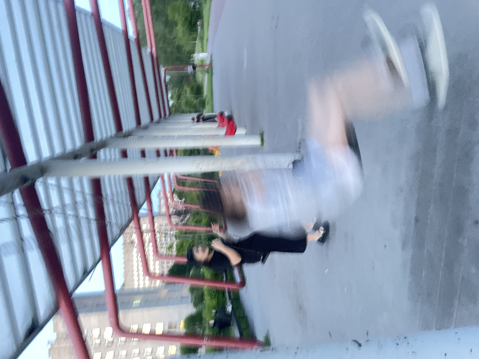The image depicts an outdoor public swing set structure, likely in a park or recreational area, in Sinyavino, Russia. A young person wearing a black outfit and a dark hat is seated on a swing, appearing to be in motion, with their arms outstretched. Another individual, whose features are blurred due to motion, is also on a swing in the foreground, with their legs and feet visible. The swing set has a red metal frame and a corrugated metal roof supported by white posts. In the background, there are tall residential buildings with visible windows and trees. The ground is a paved surface, possibly asphalt or concrete. Other people are present in the background, appearing as indistinct figures, some seated on benches and others standing or walking. The overall scene captures a moment of activity at a public playground.