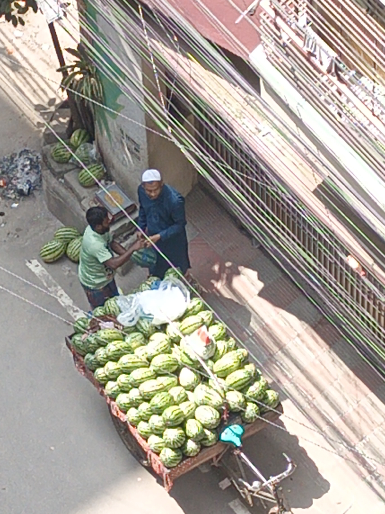 The image shows a street scene in Dhaka, Bangladesh. A watermelon vendor stands next to his bicycle cart filled with watermelons and is interacting with a customer who is paying for a purchase. The customer is wearing a traditional Muslim cap. Additional watermelons are piled on the street next to the vendor's cart and a small plant. The street is covered with overhead wires. A building is visible on the right of the image.