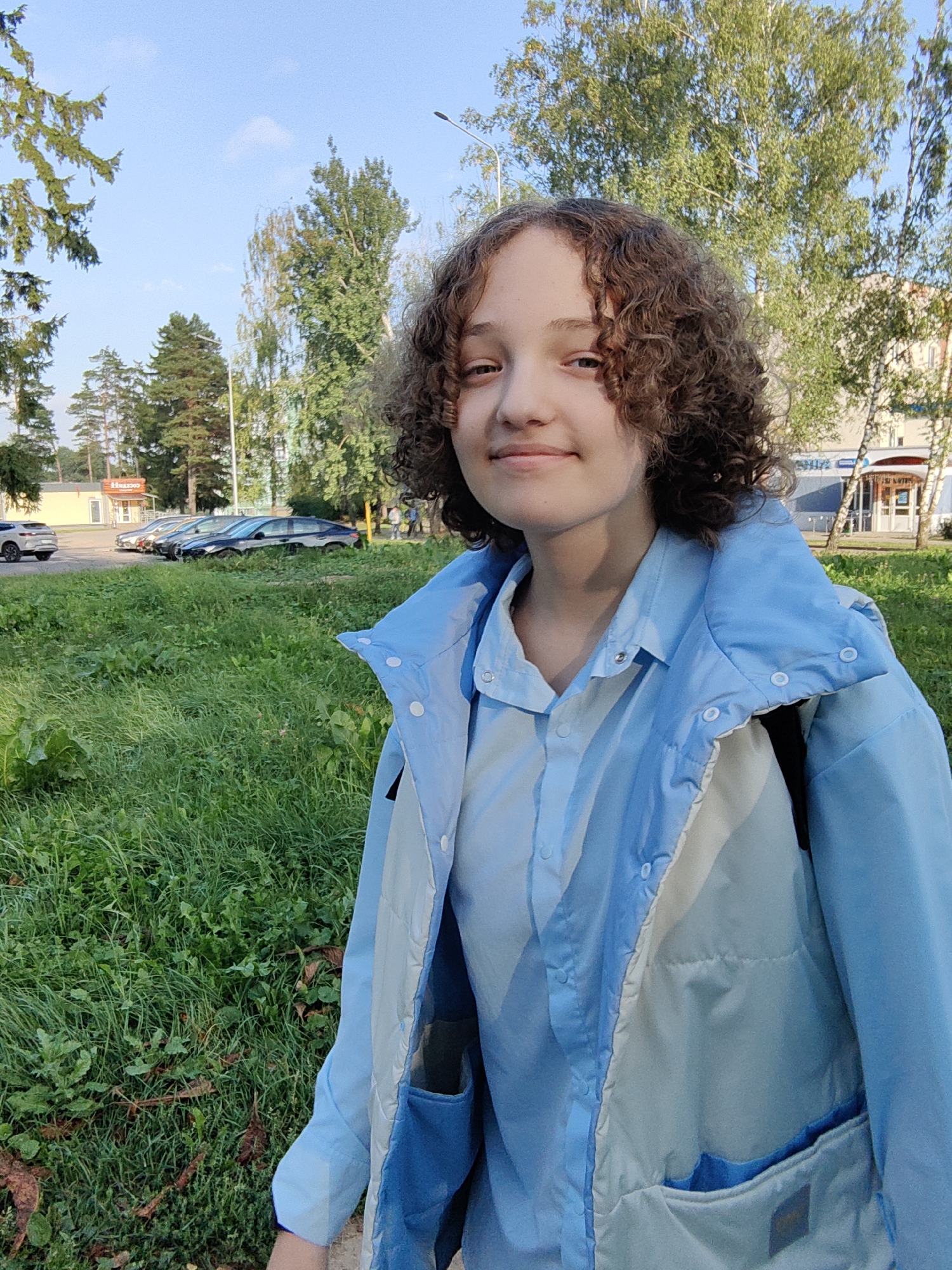 In Fanipal, Belarus, a young person with curly brown hair is captured in a medium close-up shot, smiling slightly at the camera. They are wearing a light blue button-down shirt and a light blue puffer jacket over it. The background features a grassy area with some fallen leaves, trees, and buildings. In the distance, several cars are parked on a road. The sky is clear and blue, indicating it is daytime. The overall scene appears to be an outdoor, urban or suburban setting.