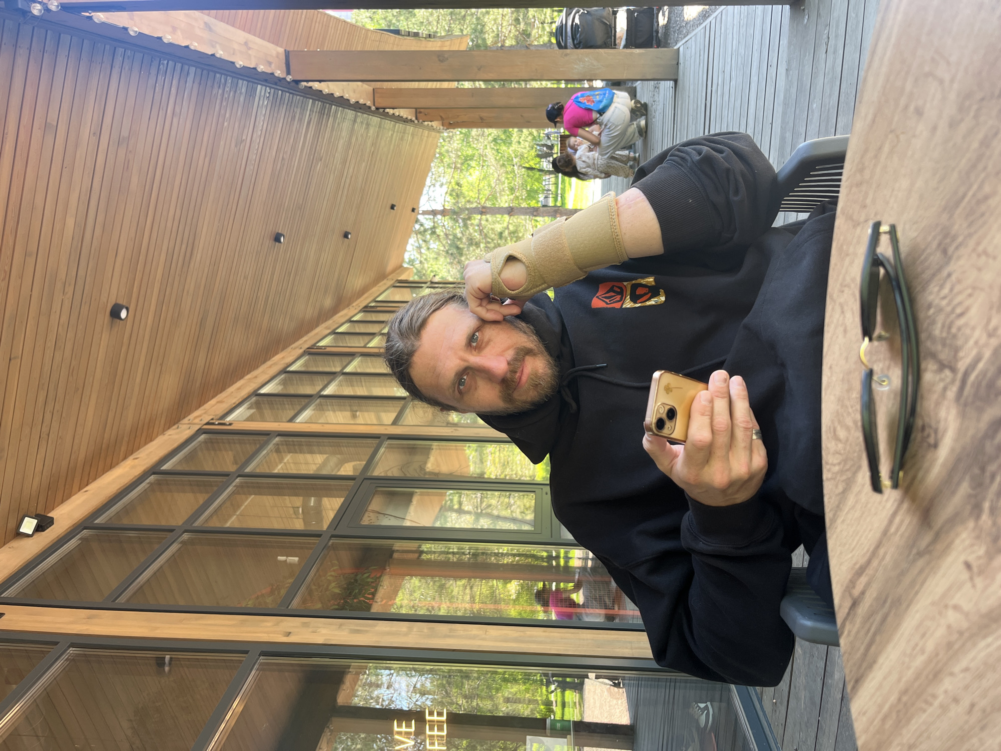 A man with a beard and long hair, wearing a black hooded sweatshirt and a light-colored wrist brace on his left arm, is seated at a wooden table outdoors. He holds a gold-colored smartphone in his right hand and looks directly at the camera with a neutral expression. On the wooden table beside him, a pair of dark-framed sunglasses rests.
The man is positioned on a wooden deck next to a building characterized by extensive vertical wooden paneling and large, multi-paned glass windows. The ceiling above the deck is also wooden and adorned with a string of small, clear lights. Through the windows and in the background, lush green trees and foliage are visible, suggesting a natural or park-like setting.
Further in the background, on the wooden deck, two individuals are seen crouched or bending over, seemingly interacting with a small child in a light-colored dress. One person wears a bright pink top. Several dark bags or backpacks are also visible on the deck behind them.
The scene takes place during bright daylight, indicative of sunny weather. Reflected on one of the large glass windows, part of a sign reads "LIVE COFFEE." The overall setting suggests a modern outdoor cafe or restaurant with a focus on natural materials, located in Krasnoyarsk, Russia.