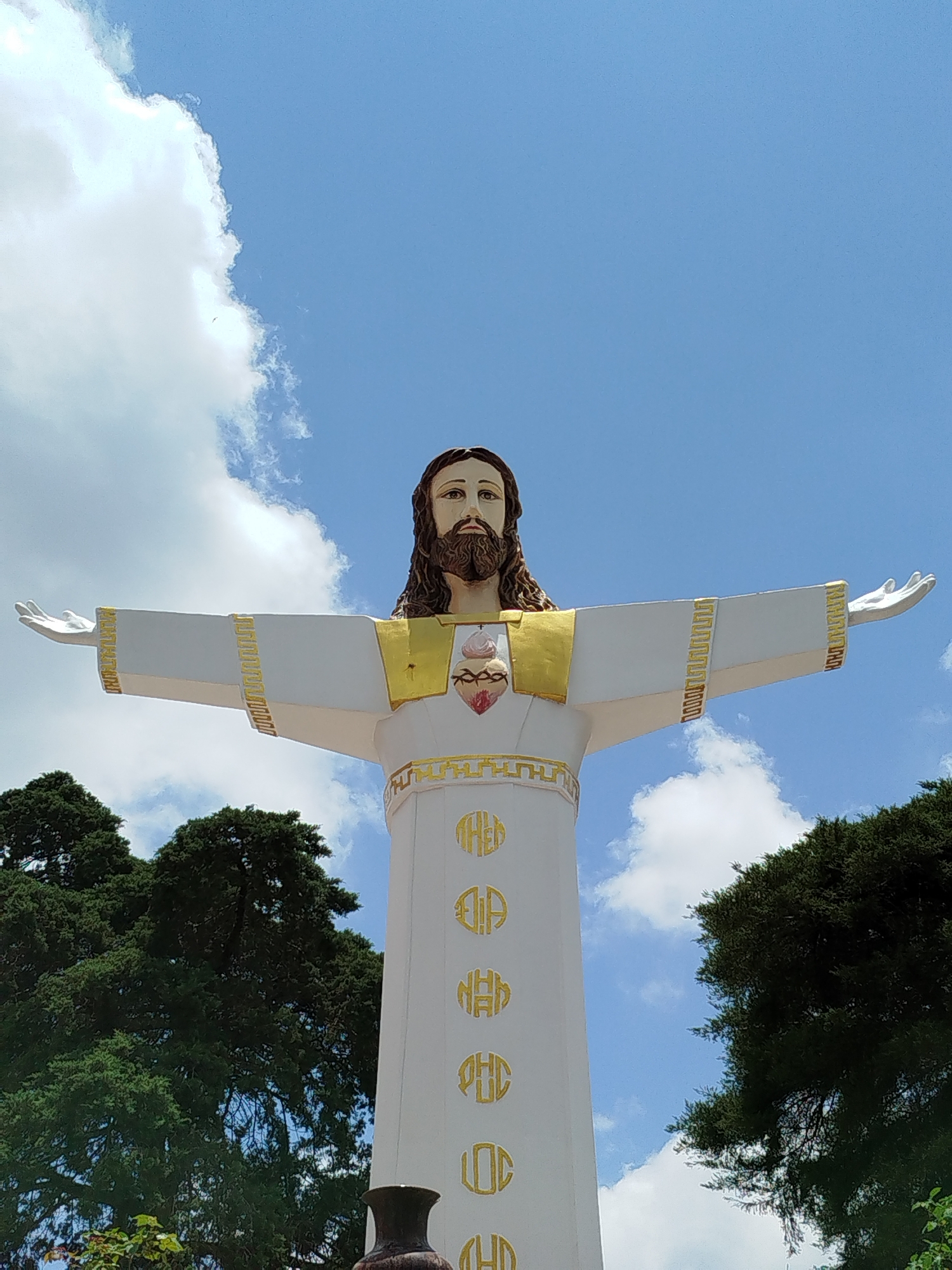 A large, towering statue of Jesus Christ is depicted in a low-angle shot against a bright blue sky with scattered white clouds, suggesting clear, sunny weather during the daytime. The statue is predominantly white, with golden accents on its robe and a brown beard and hair. Its arms are outstretched, in a welcoming or blessing gesture. On the chest of the statue, the Sacred Heart symbol is visible, rendered in red and pink hues, surrounded by golden flames and thorns.
The statue stands on a cylindrical pedestal or lower part of its robe, which is adorned with several circular golden motifs arranged vertically. These motifs appear to contain stylized letters or symbols, though they are not fully legible from this distance.
Dark green, dense foliage from trees frames the lower sides of the statue, extending upwards to about the height of its shoulders. A dark, urn-like object or pot is partially visible in the very bottom foreground, slightly to the left of the statue's base. The overall scene conveys a sense of grandeur and spirituality, typical of a religious landmark. This statue is located in Lâm Viên - Đà Lạt, Vietnam.