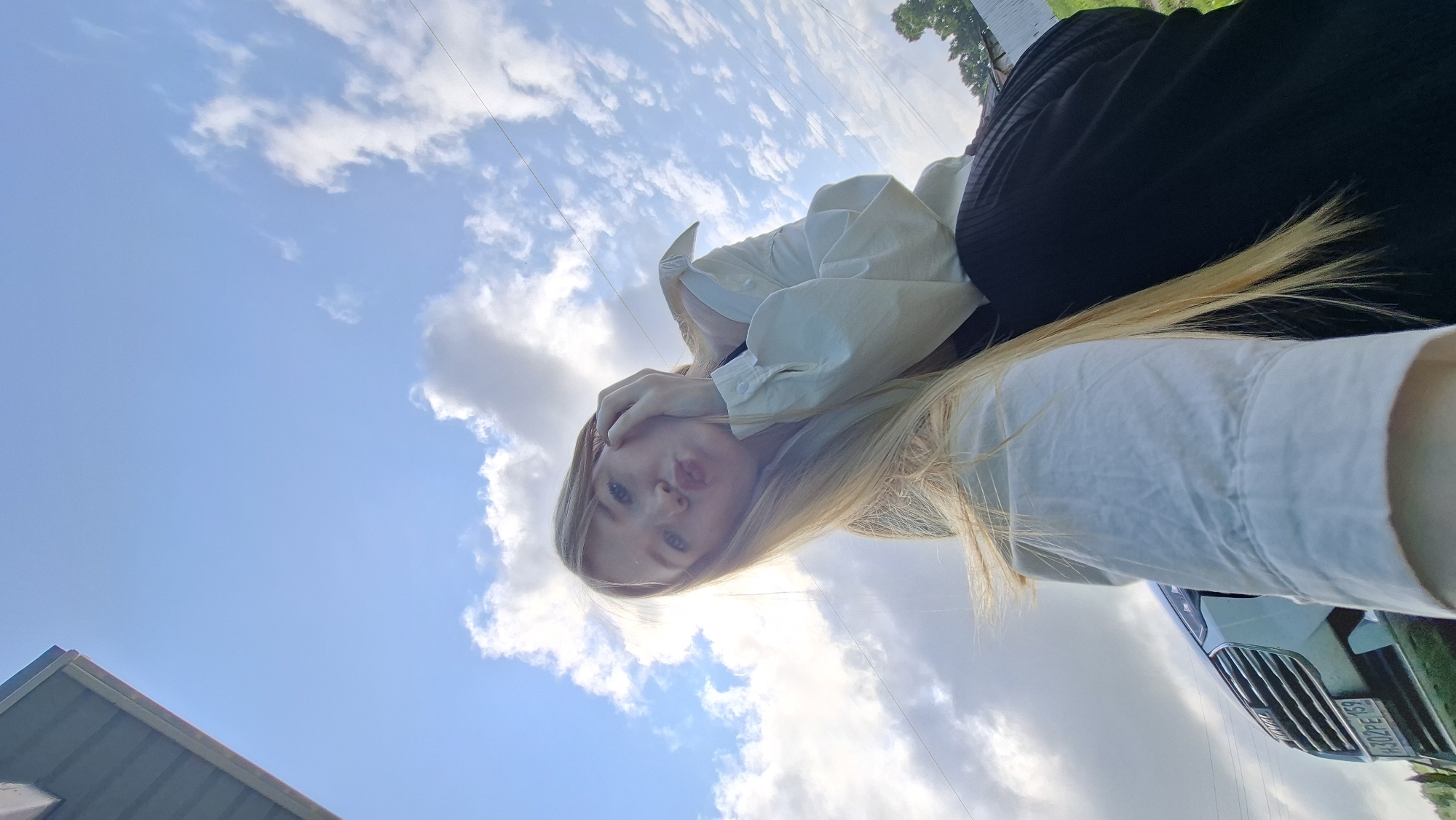 A young blonde woman is taking a low-angle selfie outdoors. She is wearing a white button-up shirt and a black skirt, and her hair is long and blonde. The sky is blue with scattered white clouds. In the background, a silver car with a license plate is visible, along with some trees and a building. The scene appears to be in a rural or suburban setting. The woman is puckering her lips and holding her hand to her cheek, suggesting a playful or affectionate pose. The overall atmosphere of the image is casual and relaxed.