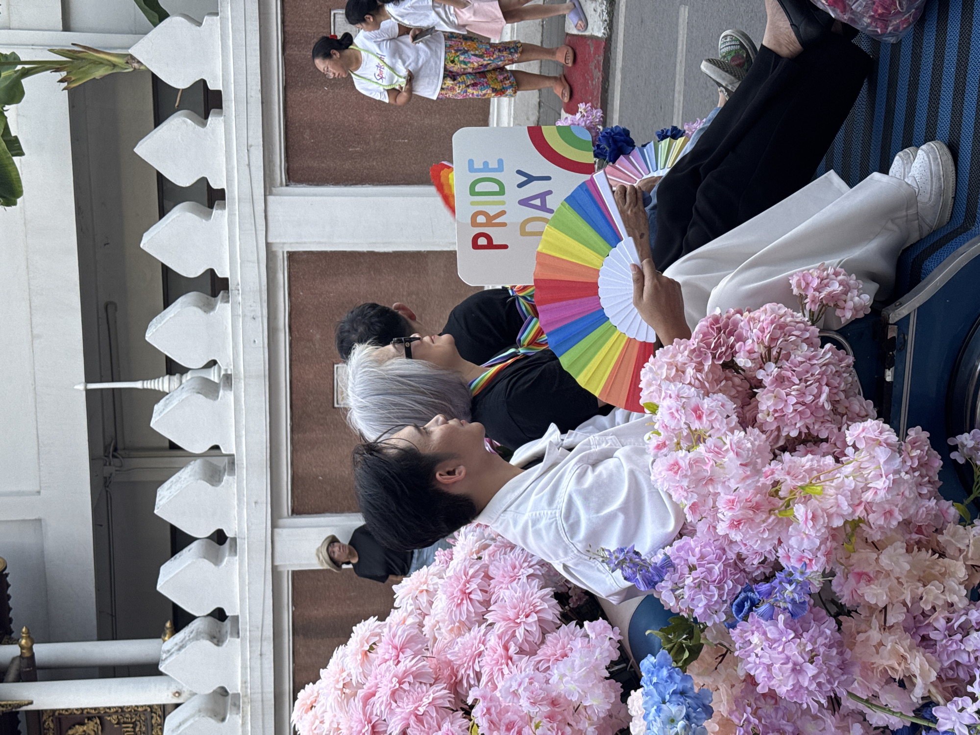 The image shows a Pride parade in Chiang Mai City Municipality, Thailand. Three people are sitting on a vehicle that is decorated with many flowers and a "Pride Day" sign. The person in the foreground is wearing a white shirt, and there are pink flowers around them. Another person is holding a rainbow fan. Two people are walking behind the vehicle. The background shows a building with white decorations.