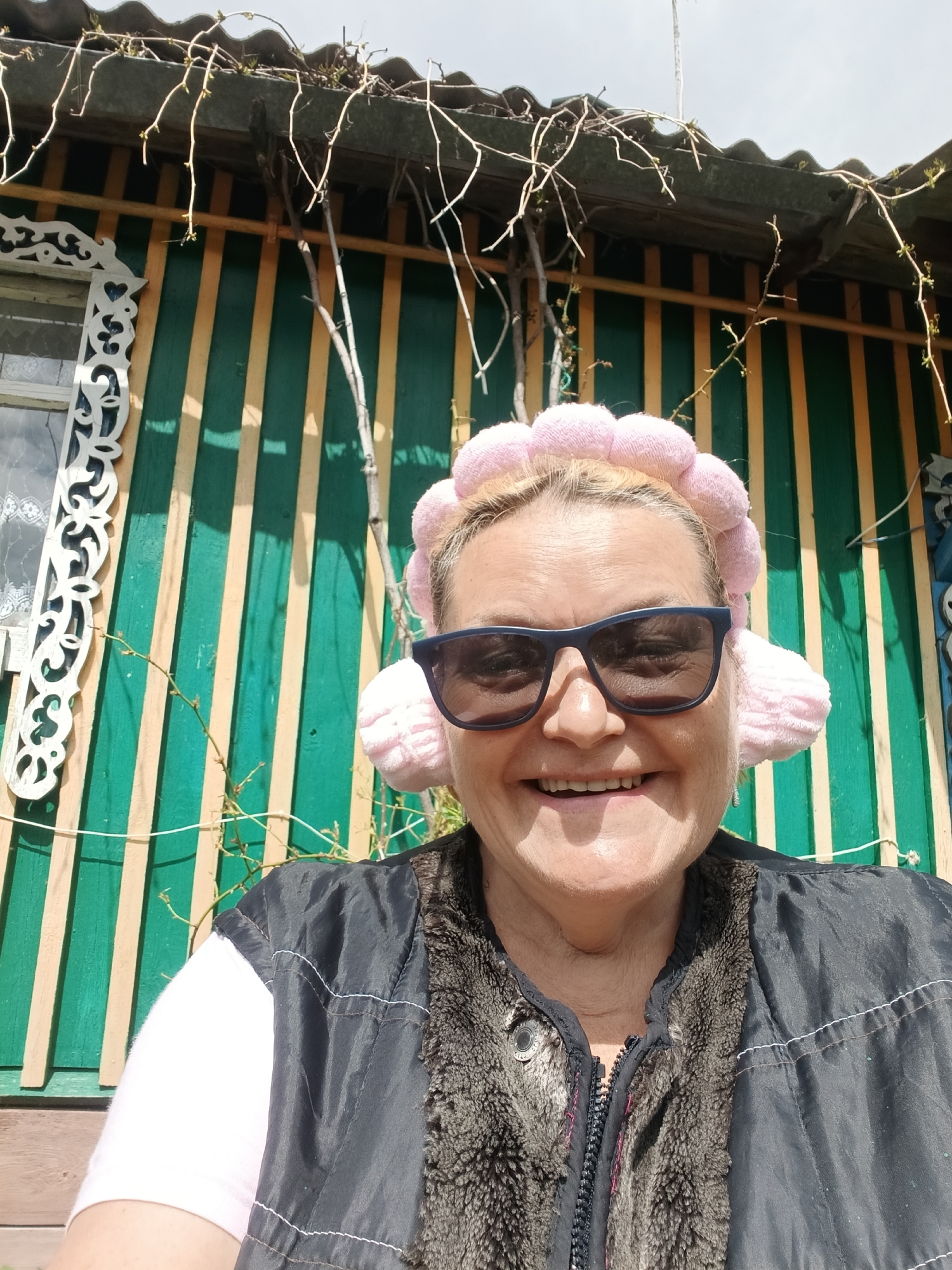 The photograph shows a middle-aged woman smiling at the camera. She wears dark sunglasses and a pink headpiece resembling a flower crown with earmuffs. She is also wearing a black vest with a fur-like collar over a pink shirt.
Behind the woman is a wall with green and yellow vertical stripes. There is also a window with decorative white trim. Bare branches are visible extending from the roof above.
Based on the information provided, the location is somewhere in the city of Красный Маяк, Russia.