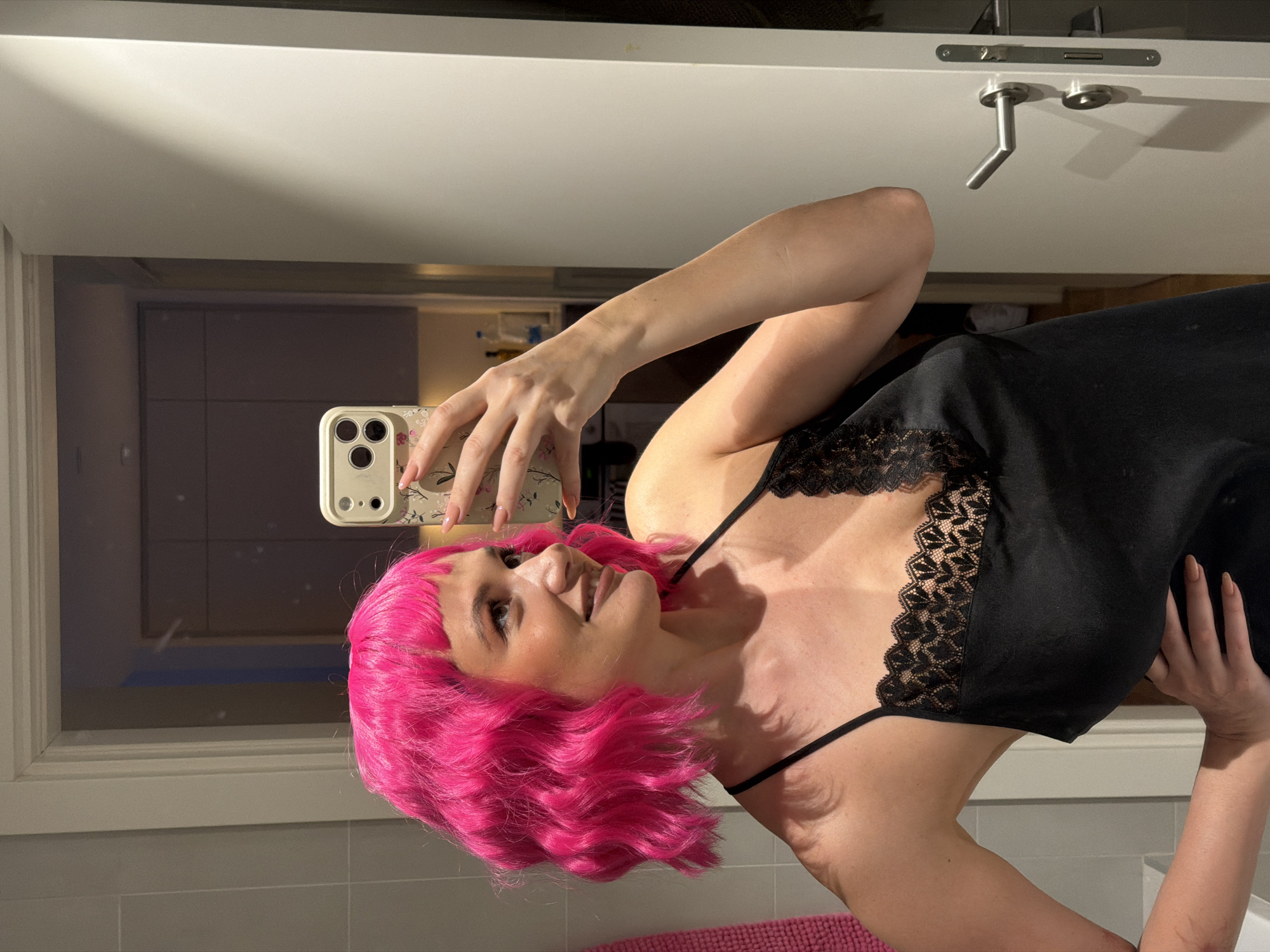 A woman with vibrant pink hair is taking a selfie in a bathroom. She is wearing a black slip dress with lace trim. Her hand is holding an iPhone with a light-colored patterned case. The bathroom has light gray tiles and white fixtures. In the reflection of the mirror, a glimpse of another room can be seen with light-colored walls and possibly kitchen cabinets. The location is not clearly depicted, but the prompt requested that it should be in Belgrade, Serbia. There is no information to confirm if this is the correct location. A pink bathmat is visible in the bottom of the frame.