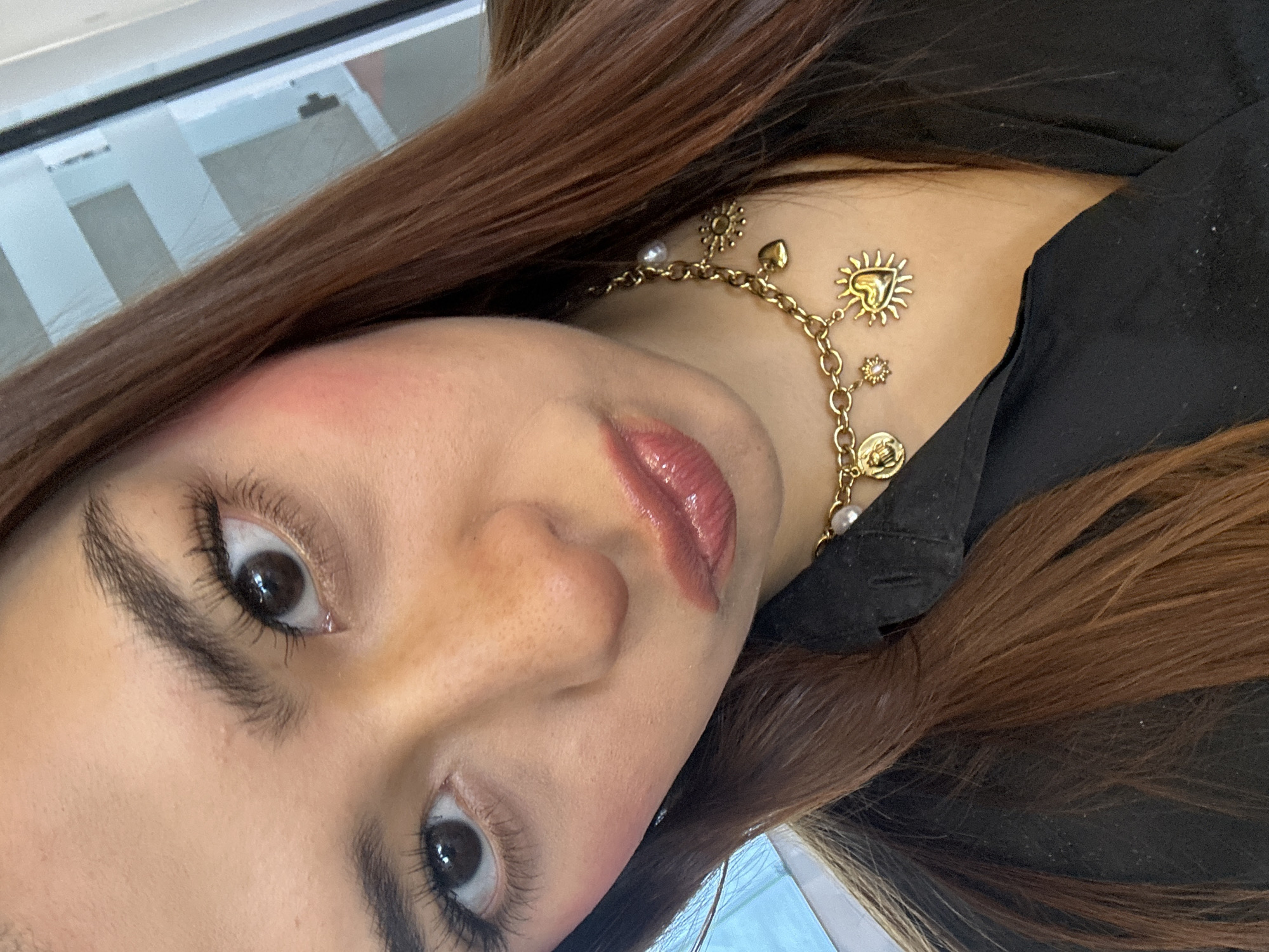 A medium close-up shot shows a person with brown hair and light skin, their face angled towards the lower left of the frame. They are wearing a gold chain necklace with various charms, including a heart within a sunburst design. The person's lips are coated with a glossy, light pink lip color. They are wearing a dark-colored top. The background is a blurred mix of white and grey, suggesting a window or reflective surface. There is no verifiable information to specify the city of San Bernardino Tlaxcalancingo, Mexico in the image.