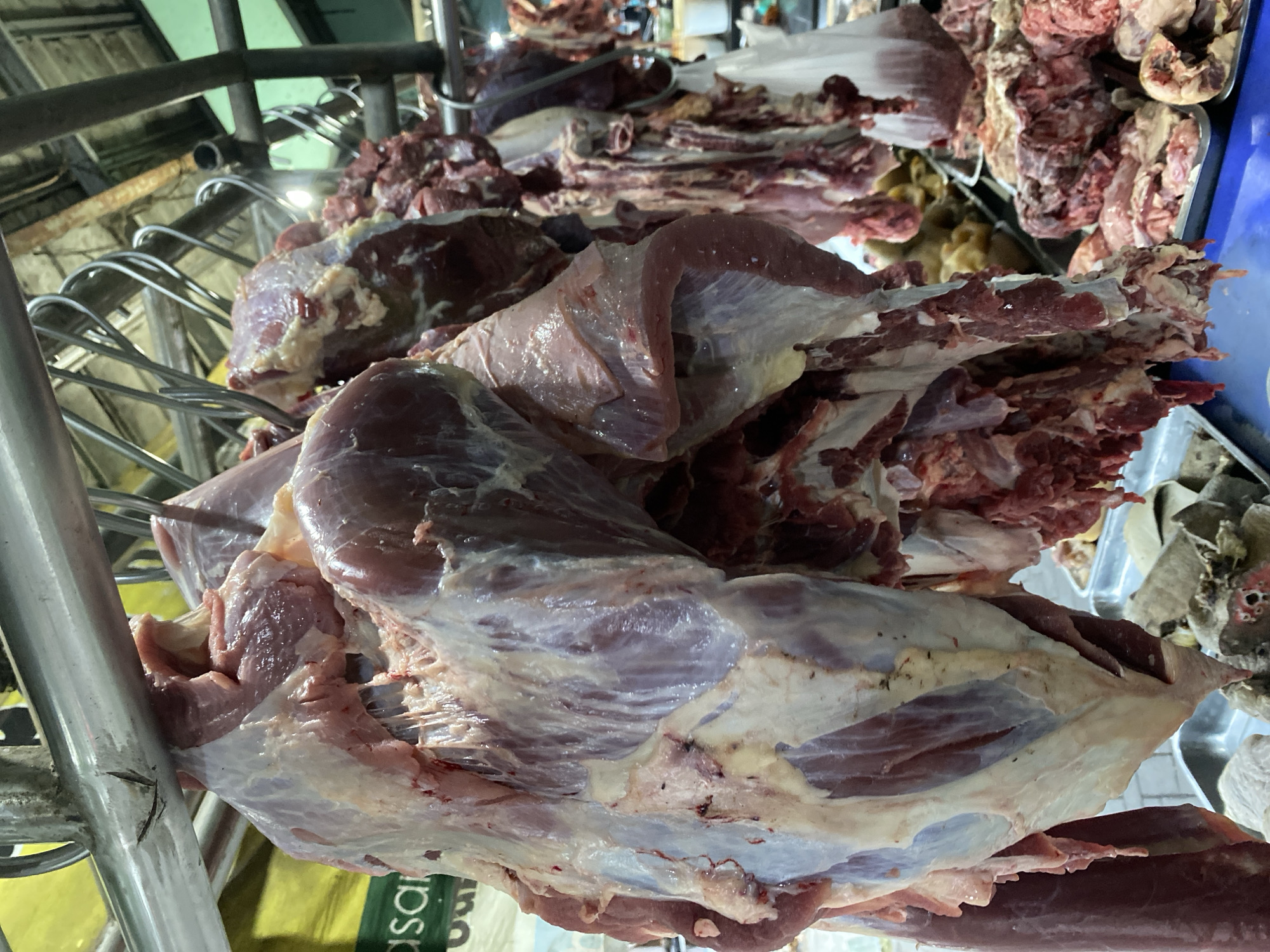 A side view of hanging meat cuts at an outdoor market stall in Ambarketawang, Indonesia. The foreground features large cuts of raw meat, some with fat and connective tissue attached, suspended from metal hooks and bars. In the background, other cuts of meat are visible, some in trays. The overall scene depicts a butcher's stall in a marketplace setting.