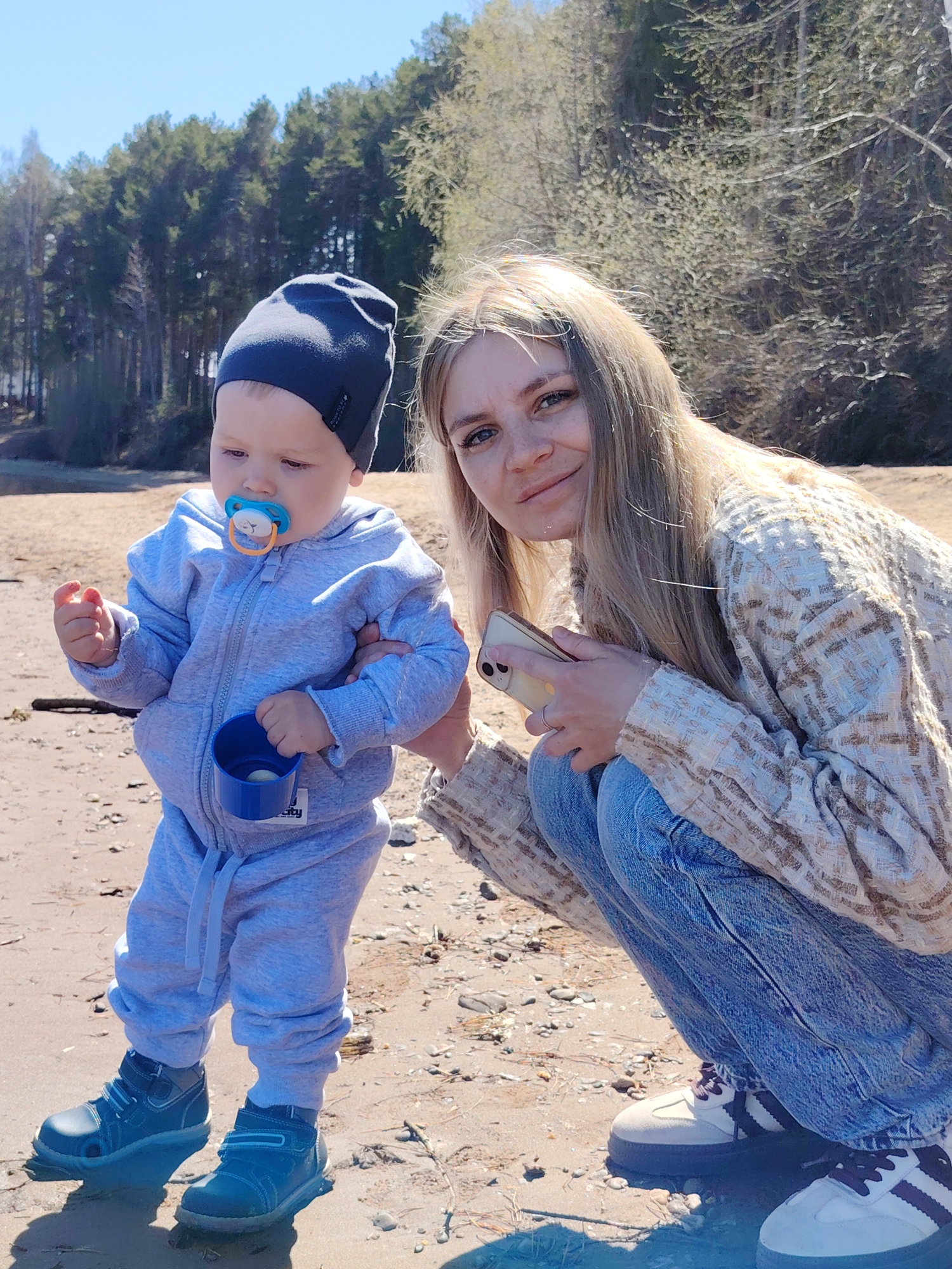 A woman and a toddler are on a sandy area, possibly a beach, near a body of water with a forested shoreline. The woman is crouching, wearing a tan and white patterned jacket, blue jeans, and white and black sneakers. She is holding a smartphone. The toddler, standing beside her, is wearing a gray jumpsuit, a dark blue beanie, and blue shoes. The child has a pacifier in his mouth and is holding a small blue cup in one hand. The background shows trees and a body of water, suggesting an outdoor, possibly recreational, setting. The location is Chaikovsky, Russia.