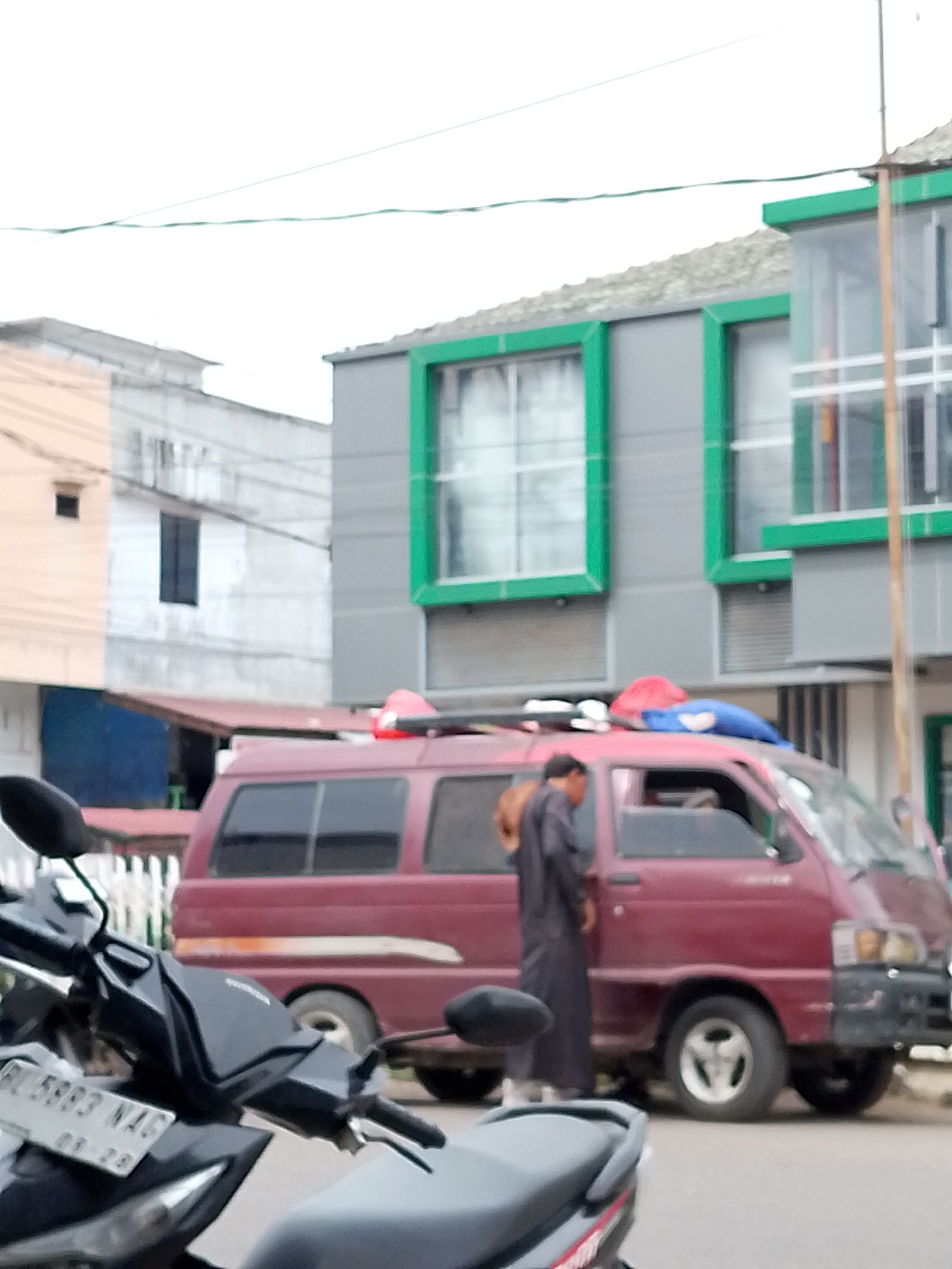 The image depicts a street scene in Lhokseumawe, Indonesia, with a maroon van as the central object. A person dressed in dark clothing, possibly a traditional robe and head covering, stands beside the driver's side door of the van. The van has a luggage rack on its roof with several items secured to it, including red and blue bags. In the foreground, a motorcycle is partially visible, showing its handlebars and part of the seat. Behind the van, a two-story building with gray siding and prominent green window frames is visible. The building has multiple windows, some of which appear to be covered with blinds. To the left of this building, another building is partially visible, appearing lighter in color with a dark rectangular window and a smaller square opening. The sky is overcast, suggesting a cloudy day. The scene appears to be an urban or semi-urban setting.