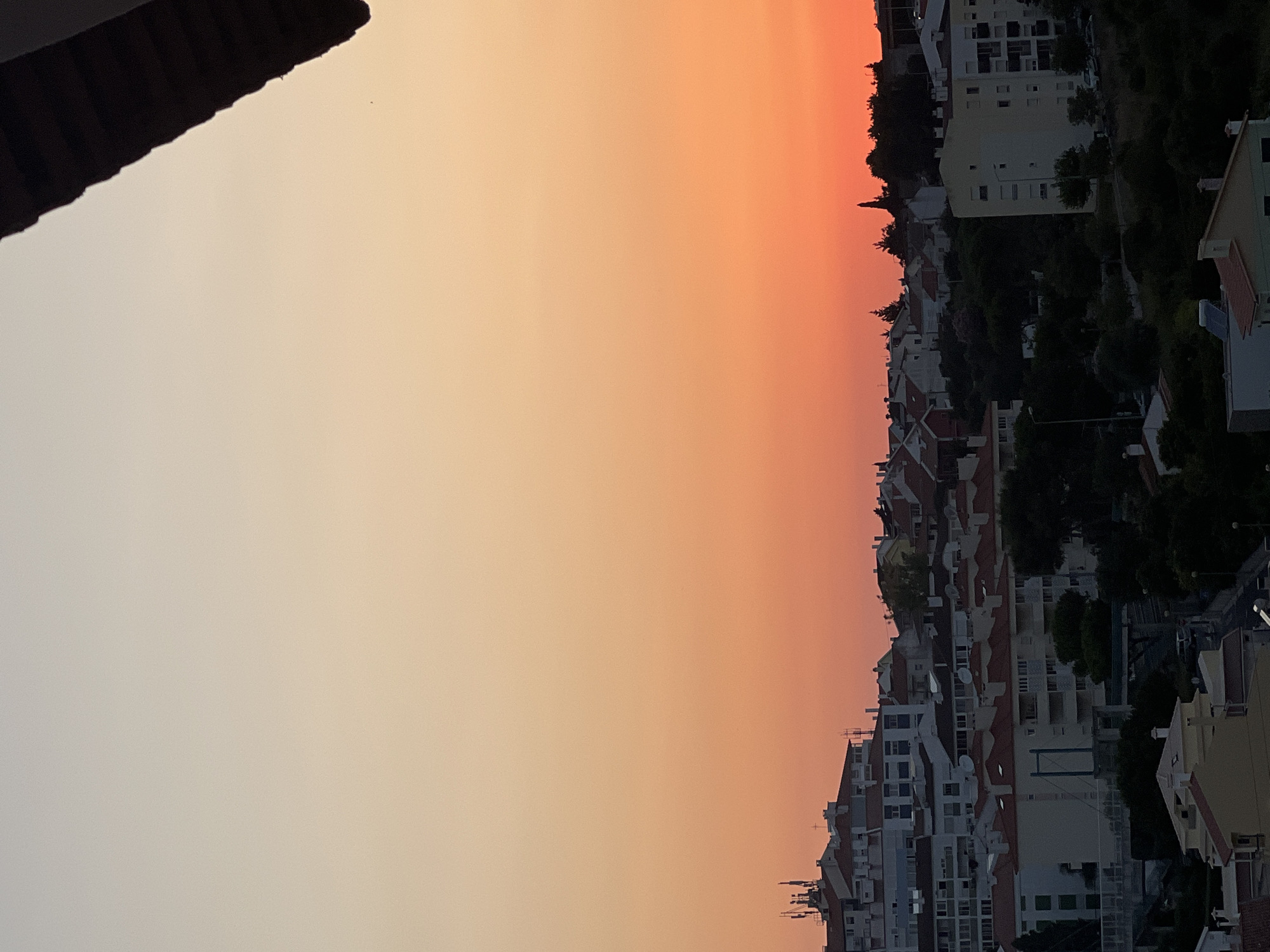 The image captures a panoramic view of a residential area in Cascais, Portugal, during sunset. The sky displays a gradient of colors, transitioning from a pale yellow at the top to a vibrant orange and red closer to the horizon. A dark, overhanging roof structure partially obscures the upper right corner of the image.
Below the sky, a densely populated hillside is covered with buildings of varying heights and architectural styles, characterized by white and cream-colored facades and red-tiled roofs. Several buildings feature balconies and windows, some illuminated, suggesting the presence of inhabitants. Antennas and communication towers are visible on some of the taller structures, indicating urban infrastructure.
The buildings are interspersed with lush green trees and vegetation, creating a contrast between the urban landscape and nature. Shadows lengthen across the scene as the sun dips below the horizon, casting a warm glow over the cityscape. The overall impression is one of a tranquil evening in a Mediterranean town.