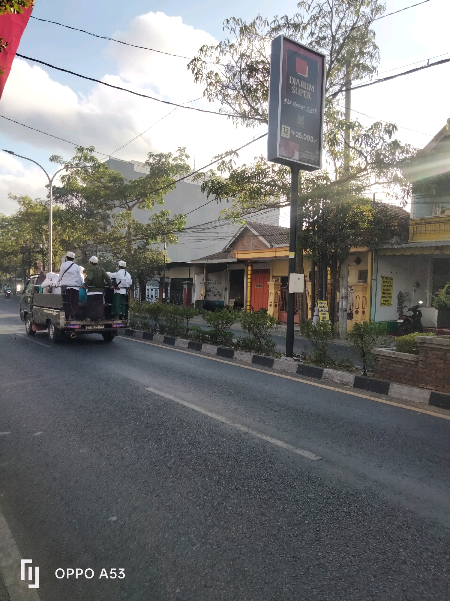 In Kelutan, Indonesia, a utility vehicle is seen driving on a road. The vehicle, a pick-up truck, is loaded with several people and items. Three individuals, dressed in white shirts and head coverings, stand in the bed of the truck. They appear to be carrying white sacks. The truck is in motion, traveling down a paved road with a striped curb on the right side. In the background, there are several buildings with residential architecture, trees, and a sign for "DJARUM SUPER". The sky is partly cloudy, with the sun casting shadows. The scene depicts a moment of transportation and activity in an Indonesian town.