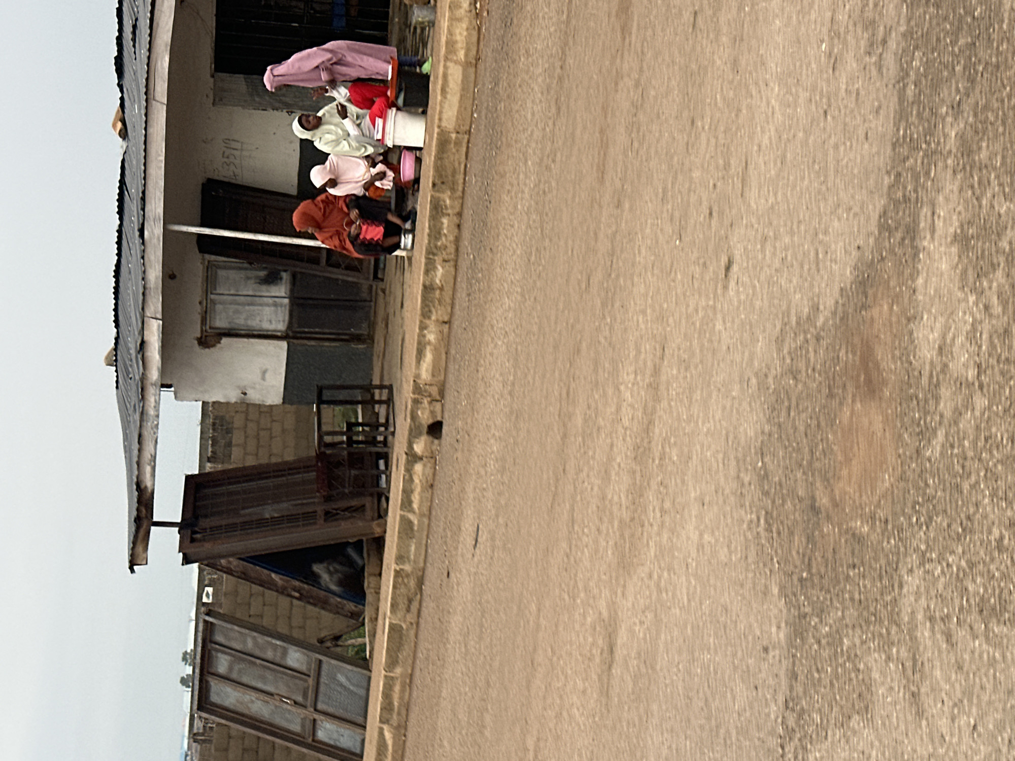 In Bukuru, Nigeria, a group of four women are gathered on a sidewalk in front of a weathered building with a corrugated metal roof. One woman, wearing a pink hijab and a long pink dress, stands and appears to be talking to the others. Three women are seated. The woman on the far left wears an orange hijab and a dark skirt. Next to her, a woman in a light pink hijab sits. The third seated woman wears a white hijab and a light-colored garment. In front of the seated women are various objects, including a pink basin, a white bucket, and some red material. To the left of the building, several large, weathered metal frames, possibly doors or windows, lean against a brick wall. A small, dark metal table and chairs are also visible near the building entrance. The street in the foreground is paved with asphalt and shows signs of wear. The sky is overcast.