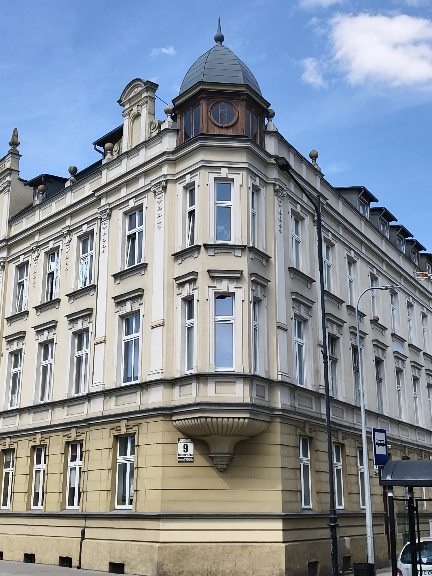 The image shows a corner view of a multi-story building with a distinct architectural style, likely dating from the late 19th or early 20th century. The building is painted in light beige and cream colors, with intricate decorative moldings around the windows and along the roofline. A prominent feature is a domed turret on the corner of the building, topped with a spire. The windows are tall and narrow, many with white frames.
The building is located at the intersection of two streets, as indicated by the street sign displaying "9" and "Kopernika". This address suggests the location is in Tczew, Poland, as Kopernika is a common street name in Polish cities and "9" is likely a building number.
In the foreground, there is a sidewalk, a dark lamppost, and part of a bus stop shelter. The sky is blue with scattered white clouds. The overall scene depicts an urban environment with a blend of historical architecture and modern infrastructure. There are no people visible in the image.
