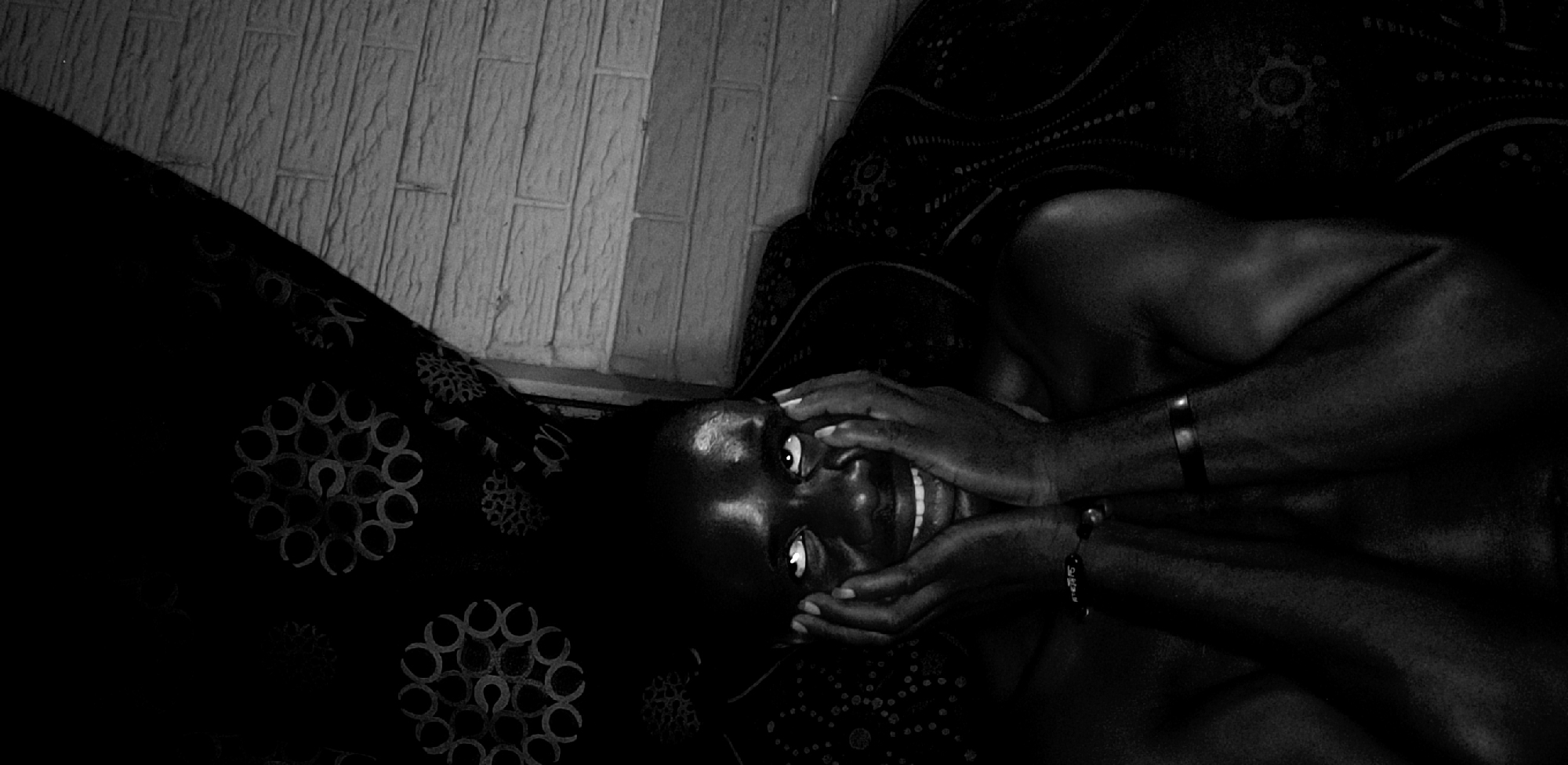 The black and white image shows a person lying on what appears to be a patterned sofa, with their head resting near a wall. The person has dark skin, and they are looking directly at the camera with their hands framing their face. They appear to be smiling. The sofa has a floral or circular patterned fabric. The wall behind them is made of rectangular blocks, possibly bricks or tiles. There is no further location information available from the image alone to specify a precise location in Narayi, Nigeria.
