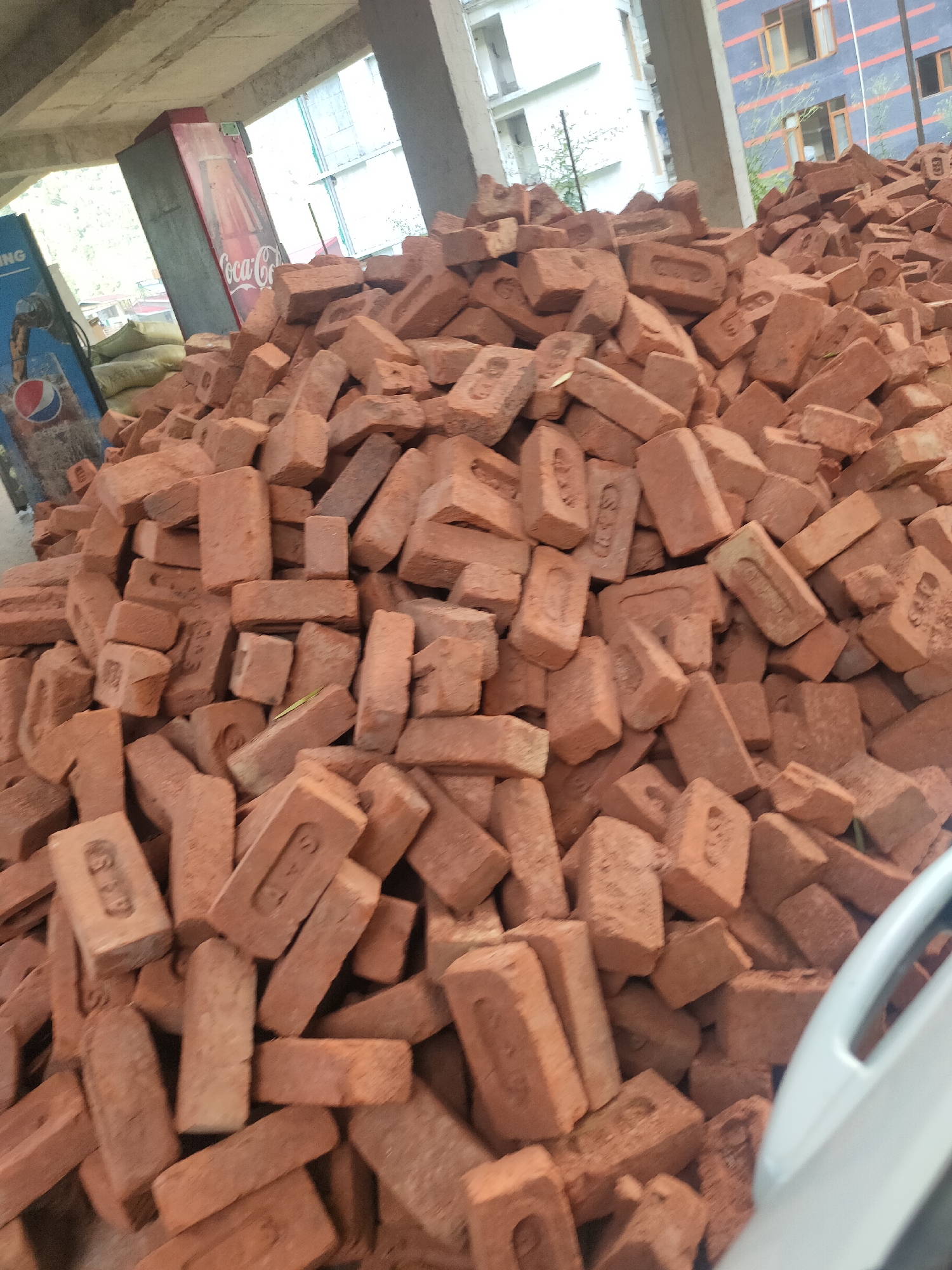 A large pile of red bricks is stacked outdoors, likely at a construction site. Behind the bricks, a Coca-Cola vending machine is visible, along with some sandbags. A building with multiple floors and windows stands in the background. The scene appears to be in the city of Siumsa, India, based on the provided information. No people are visible in the image. The lighting suggests it is daytime.