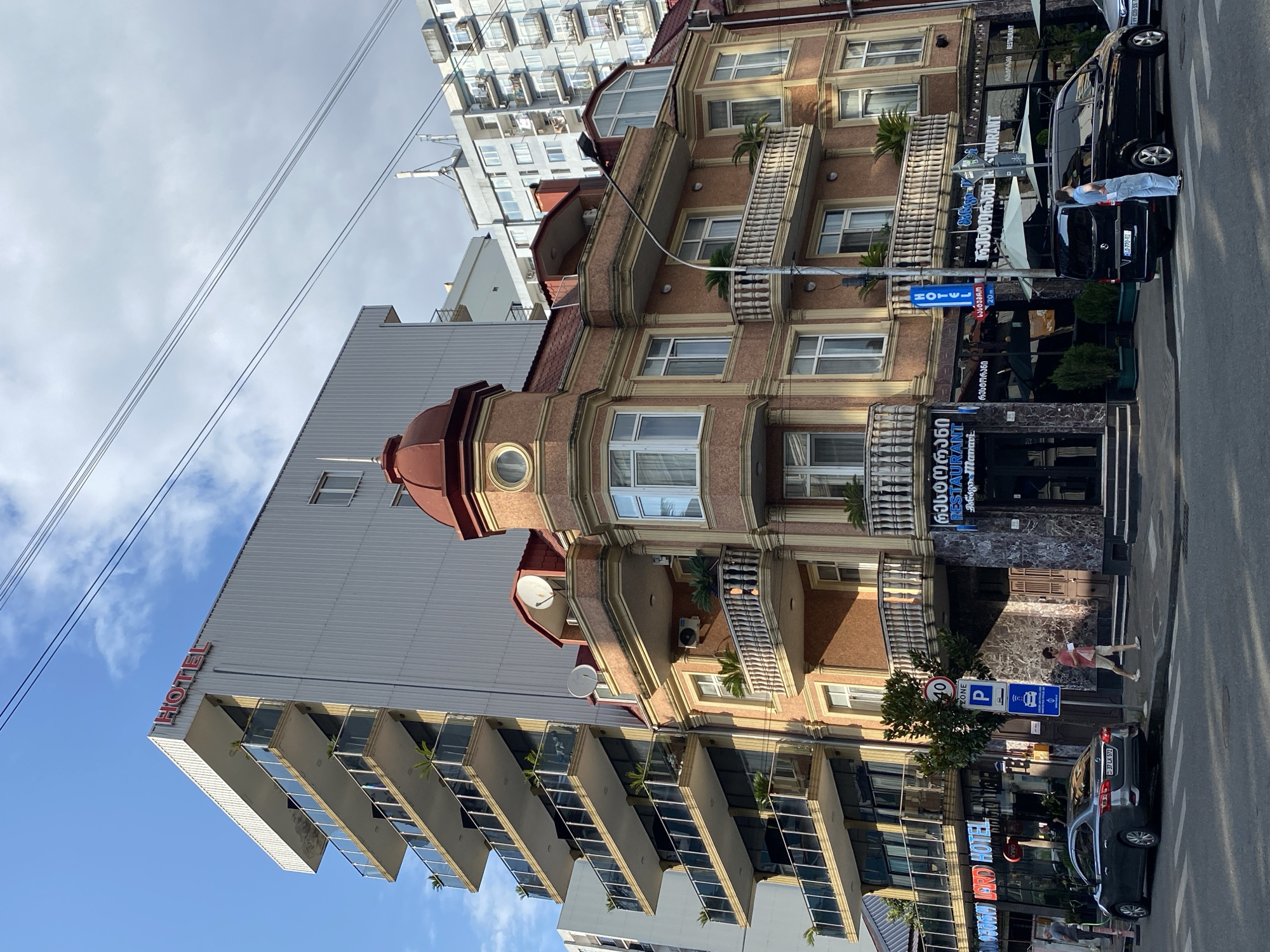 The image depicts a street scene in Batumi, Georgia, featuring a mix of architectural styles. On the left, a tall, modern building with multiple balconies and glass railings is prominently visible, with a red "HOTEL" sign at its apex. To its right, a more ornate, older building with a red domed roof and arched windows stands out. This building has balconies with decorative railings and is adorned with elaborate trim. The ground floor of both buildings appears to house businesses, including a restaurant with large windows and outdoor seating areas. Several cars are parked on the street, including a dark sedan and a black minivan. People are seen walking on the sidewalk. The sky is blue with some clouds, and electrical wires crisscross overhead, indicating an urban setting. Traffic signs, including a speed limit sign and a parking sign, are visible along the sidewalk. The overall impression is a bustling urban environment where contemporary and historical architecture coexist.