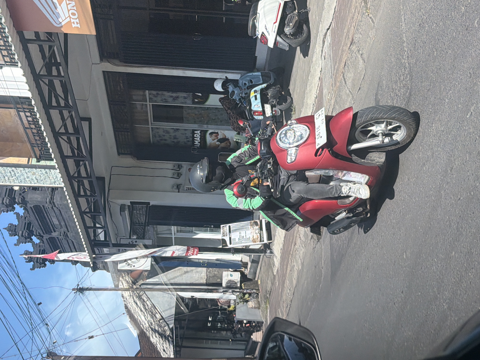 The image depicts a street scene in North Kuta, Indonesia, likely during daytime, under a clear, sunny sky. The primary subject is a person riding a red Honda scooter, positioned centrally in the frame. The rider wears a black full-face helmet, a distinctive green and black jacket (possibly a uniform for a ride-hailing service), dark pants, and white sneakers. The scooter's license plate, "P 6616 GI," with an expiration date "02-29," is clearly visible on the front fender.
The street is paved with gray asphalt, and a curb runs along the left side. To the left, a row of commercial buildings lines the street. One building features an orange awning with "HONDA" partially visible. Another shop has a prominent sign that reads "AMARA BEAUTY & SPA," featuring images of people. Below it, another sign has the word "MOTOR." Traditional Balinese architectural elements, such as ornate stone carvings, are visible on some structures. Several other scooters are parked along the street, including a light blue/gray scooter and a white Vespa-style scooter.
Overhead, a complex network of tangled power lines crisscrosses against the bright blue sky, with a red and white flag or pennant also visible. The scene captures a typical bustling street environment in a Southeast Asian urban area.