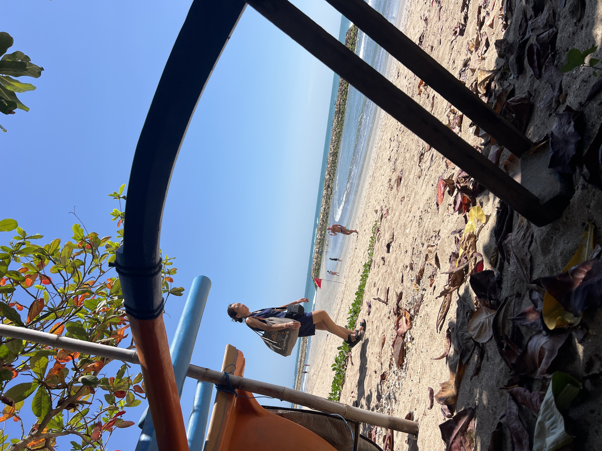 An adult female walks on the sandy shore of Kuta Beach, Indonesia, as a boat rests on the sand. The woman, wearing a dark blue dress and sandals, carries a large tote bag on her left shoulder and a smaller black bag on her right. She is positioned in the foreground, looking towards the right of the frame. Behind her, the beach extends to the ocean, with waves gently breaking on the shore. A stone jetty or breakwater stretches across the sea in the mid-ground. Scattered leaves cover the sand in the foreground, with branches and parts of the boat obscuring portions of the scene. The sky is clear and blue.