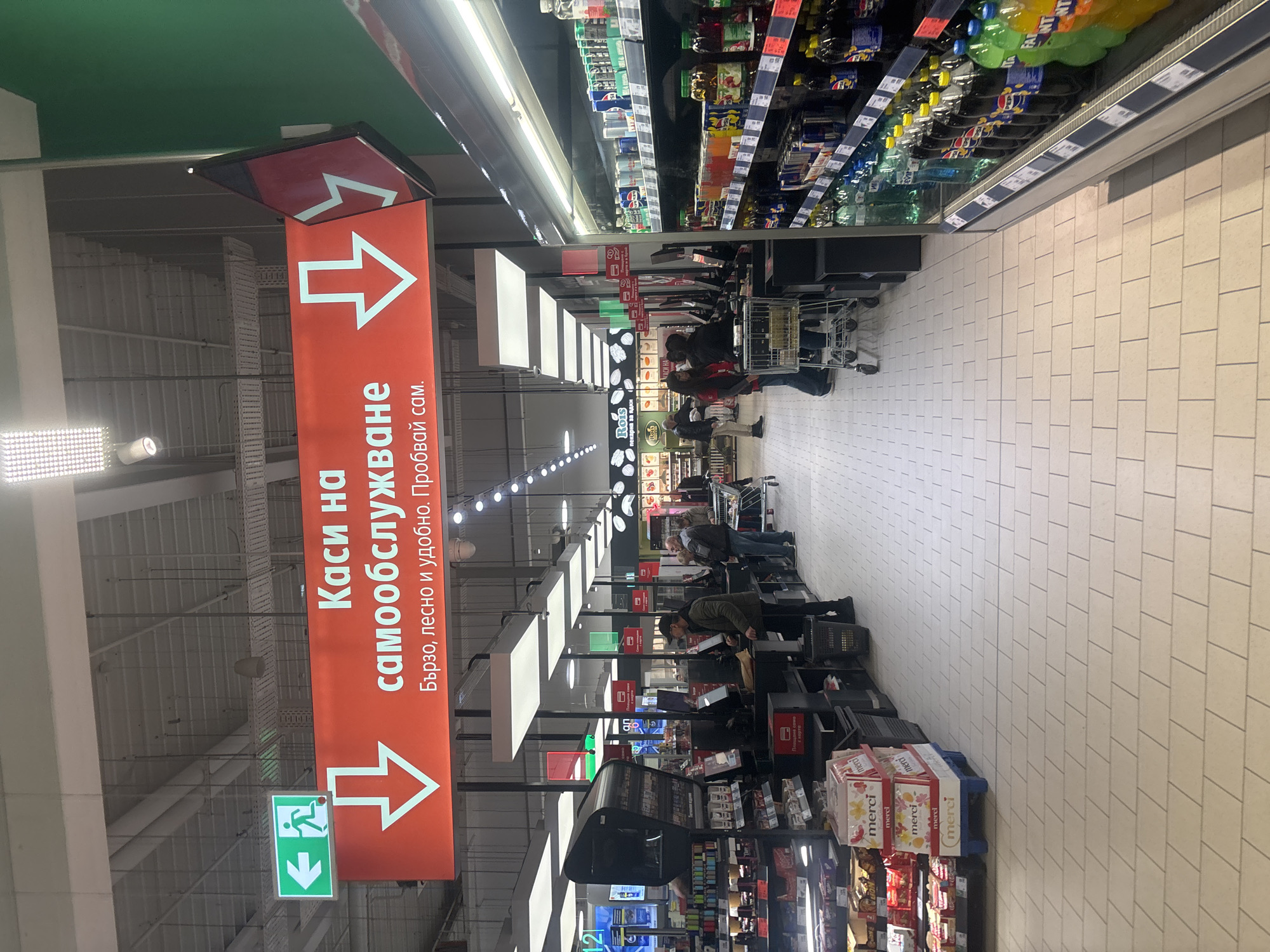 A sideways, indoor shot shows an aisle in a self-checkout area in a supermarket in Plovdiv, Bulgaria. At the top of the frame are green ceilings with light fixtures. Shelves with merchandise, drinks, and other goods are on the right and along the back wall. On the right side are self-checkout stations. Customers are at the checkout stations, and shopping carts are visible. An orange sign hangs from the ceiling, indicating the self-checkout area. Text on the sign reads “Каси на самообслужване. Бързо, лесно и удобно. Пробвай сам.” or "Self-checkout counters. Quick, easy and convenient. Try it yourself." Arrows point toward the checkout stations. An exit sign is below the arrow. The floor is tiled.