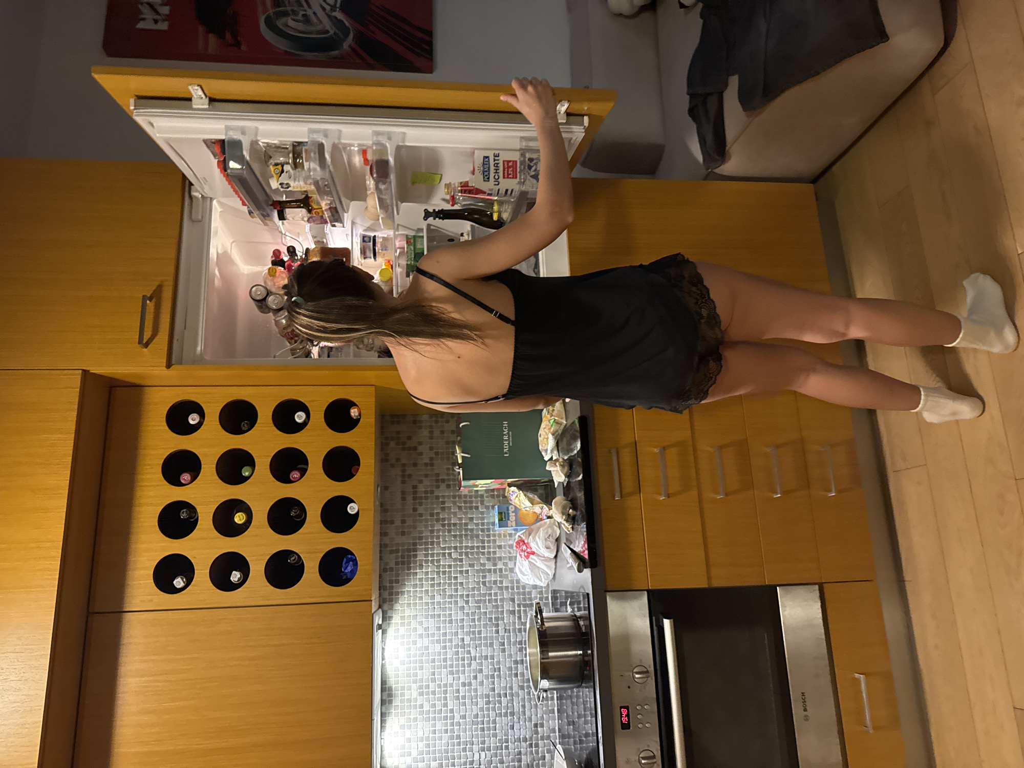 A woman wearing a black nightgown and white socks stands in a kitchen with light wood cabinets and a grey tiled backsplash. She is opening a refrigerator to her left. A wine rack built into the cabinets contains bottles of wine. A gas stovetop and oven are adjacent to the counter where a pot sits. The kitchen appears to be in Warsaw, Poland, as indicated by the "LAUDERACH" bag on the counter, a Swiss chocolate brand that has locations in Warsaw. A poster is hanging above the refrigerator. A light-colored sofa sits to the right of the cabinets. The floor is made of wood.