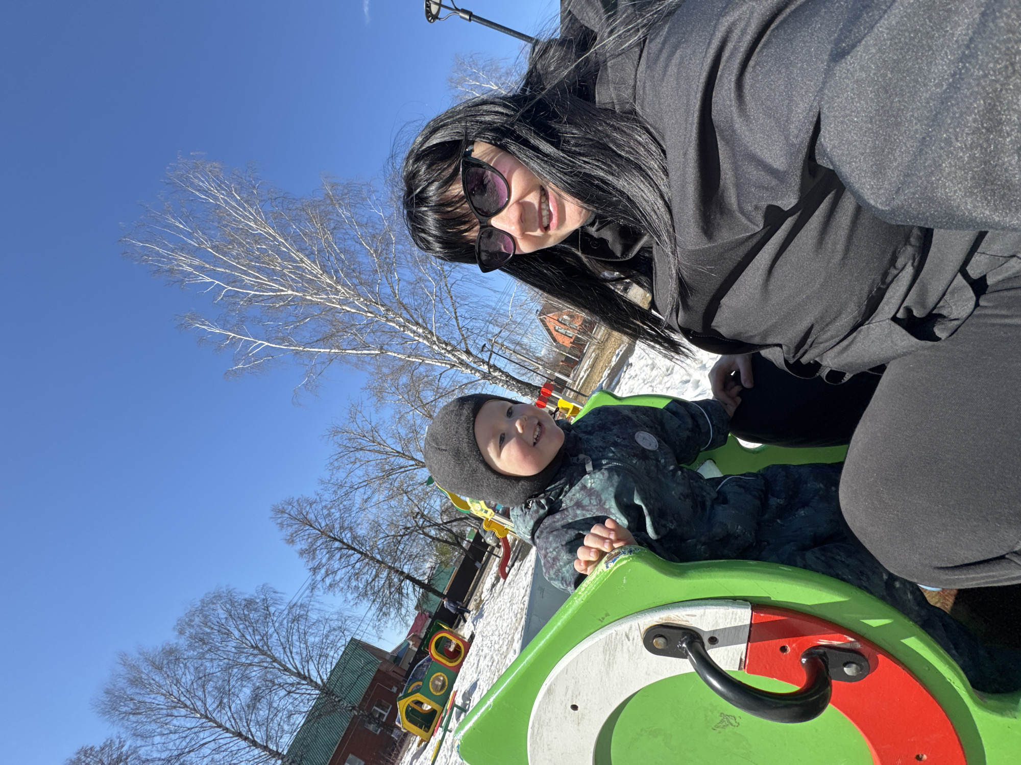 The image captures a sunny day at a playground, likely in Zav'yalovo, Russia. A woman with dark hair and sunglasses leans into the frame, smiling, while a young child wearing a dark winter hat and patterned snowsuit sits on a green and red playground carousel. Patches of snow cover the ground near a row of colorful play structures. The sky is a clear, bright blue, and bare trees are visible in the background, along with a brick building with a green roof. The woman and child appear to be interacting on the playground equipment.