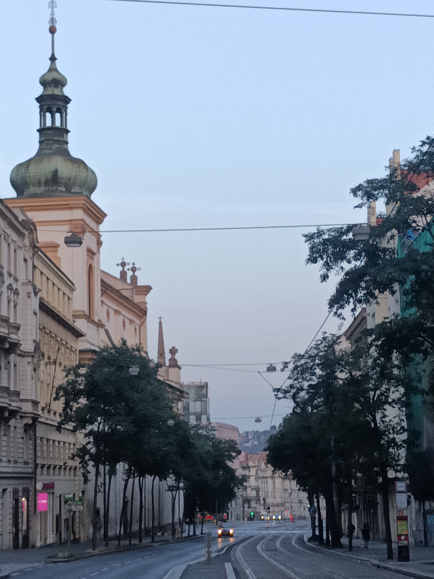 The image depicts a street scene in Prague, Czechia, likely during twilight or early morning. A tram track runs down the center of the wet asphalt road, bordered by buildings on either side and trees lining the sidewalk. On the left, a church steeple with a green dome is visible, partially obscured by the facade of a cream-colored building. The building on the right is multi-colored, with a green section on its upper right. The sky is overcast and pale blue. A few cars are visible in the distance, their headlights on. Two people are walking on the right sidewalk. Overhead, tram wires and streetlights are suspended across the street. The overall impression is of a quiet, urban environment with a mix of historical and modern architecture.