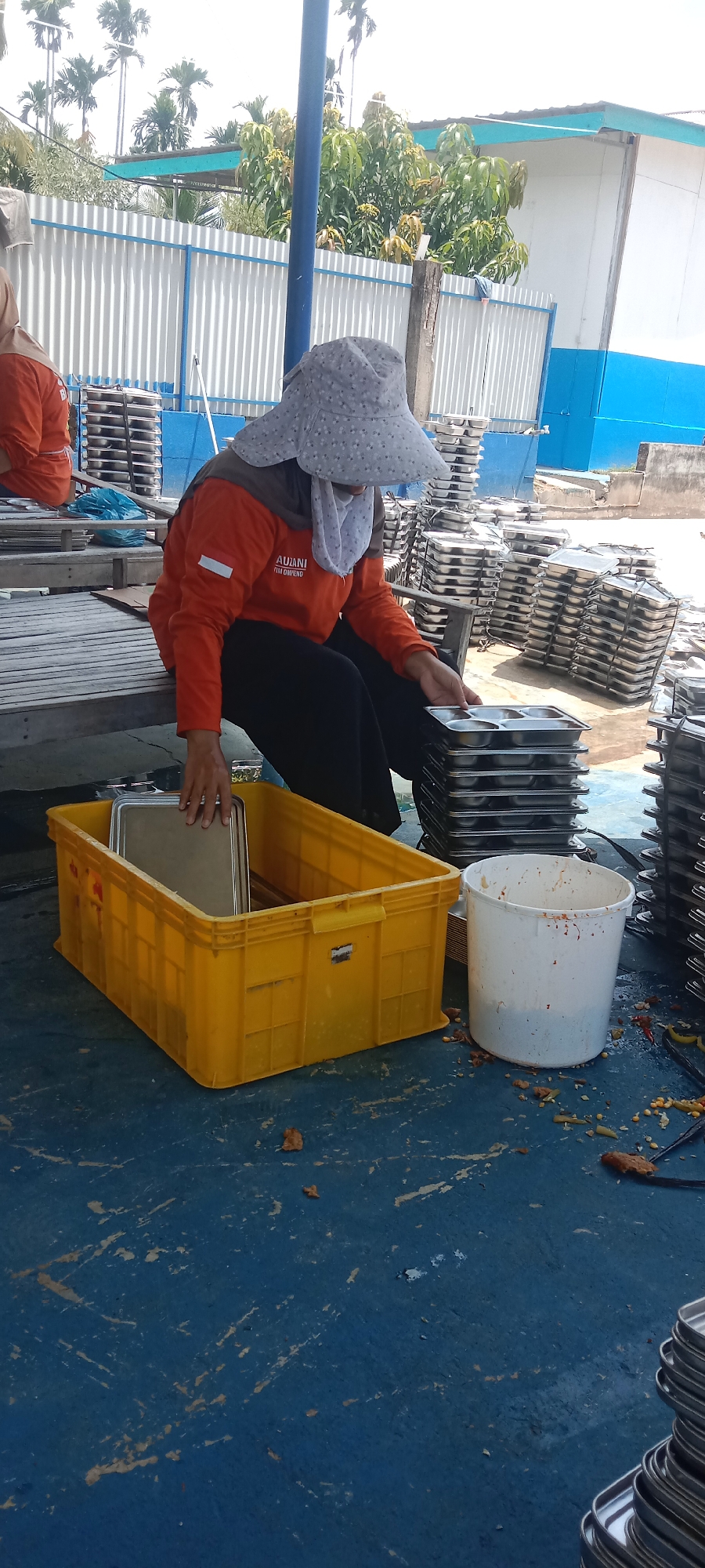In Puloblang, Indonesia, two individuals, likely workers, are engaged in an outdoor task on what appears to be an industrial site. The foreground shows a bright yellow plastic bin filled with metal trays. Beside it sits a white bucket, with remnants of food scattered on the blue concrete floor. A person wearing an orange long-sleeved shirt, dark pants, and a patterned head covering is seated, reaching into the yellow bin. Another person, partially visible on the left, also wears an orange shirt and a head covering. In the background, stacks of metal trays are piled high, and a corrugated metal fence with blue trim runs along the perimeter. Lush green trees and palm trees are visible against a bright sky, suggesting a tropical climate. The overall scene depicts a functional, possibly food processing or packaging, area.