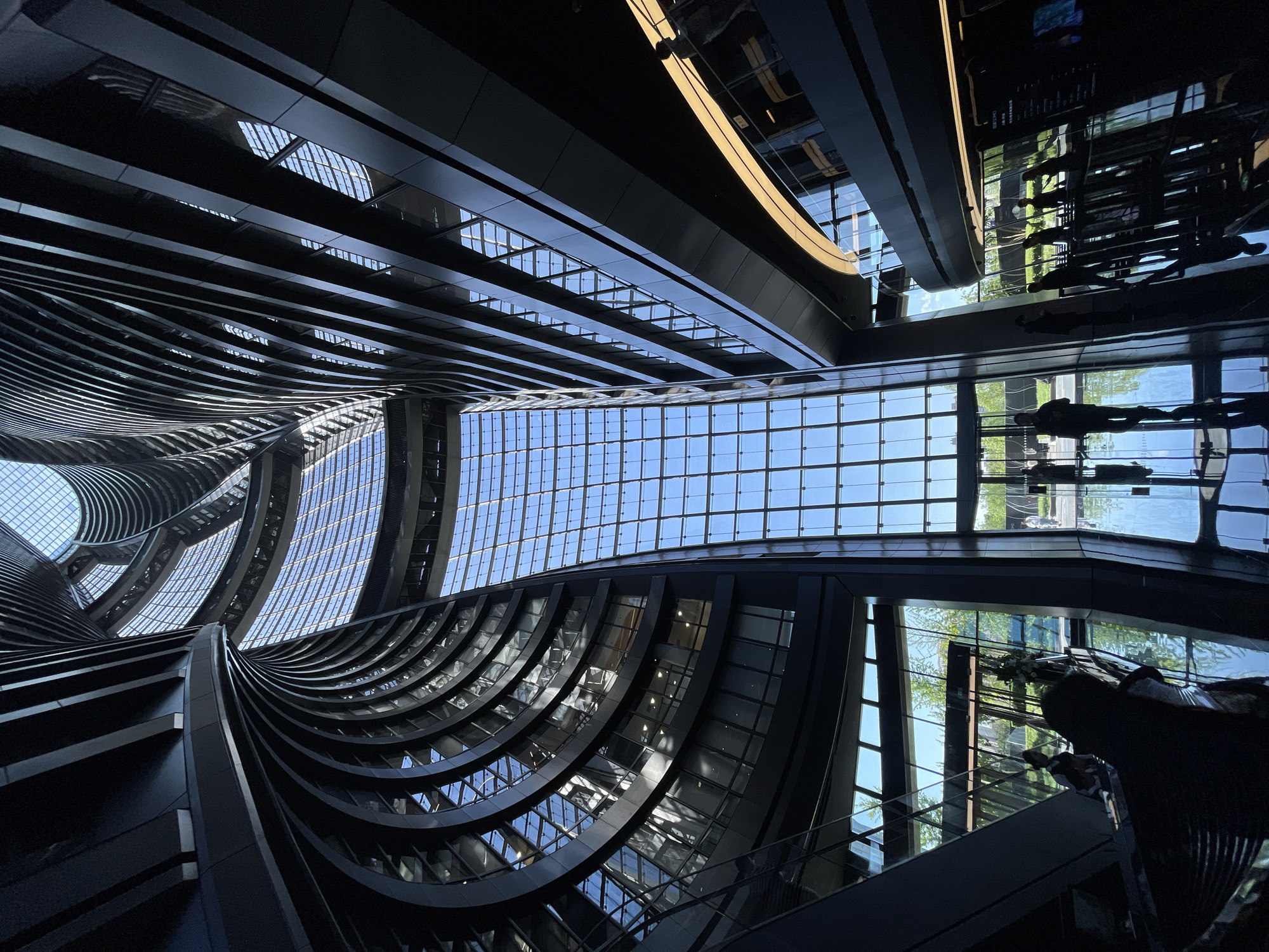 The image shows the interior of a modern building in the Fengtai District of China. The architecture is characterized by curved, black structures and a grid-like glass ceiling through which blue sky is visible. The right side of the image has mirrored glass, reflecting the exterior environment. People are visible through the glass and in the reflection of the interior of the building, which contains a statue.