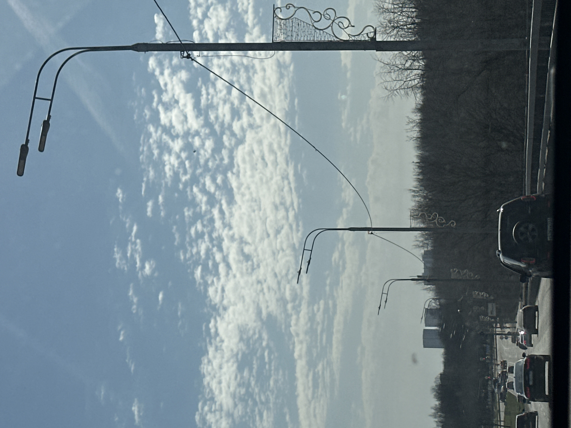 The image shows a street scene in Moscow, Russia, with several light poles lining the side of a busy road. The sky is partly cloudy with blue patches. A line of trees separates the road from buildings in the distance. Cars are seen in traffic on the road. The image was likely taken from inside a vehicle, possibly a car or bus, due to the partial view of the interior.