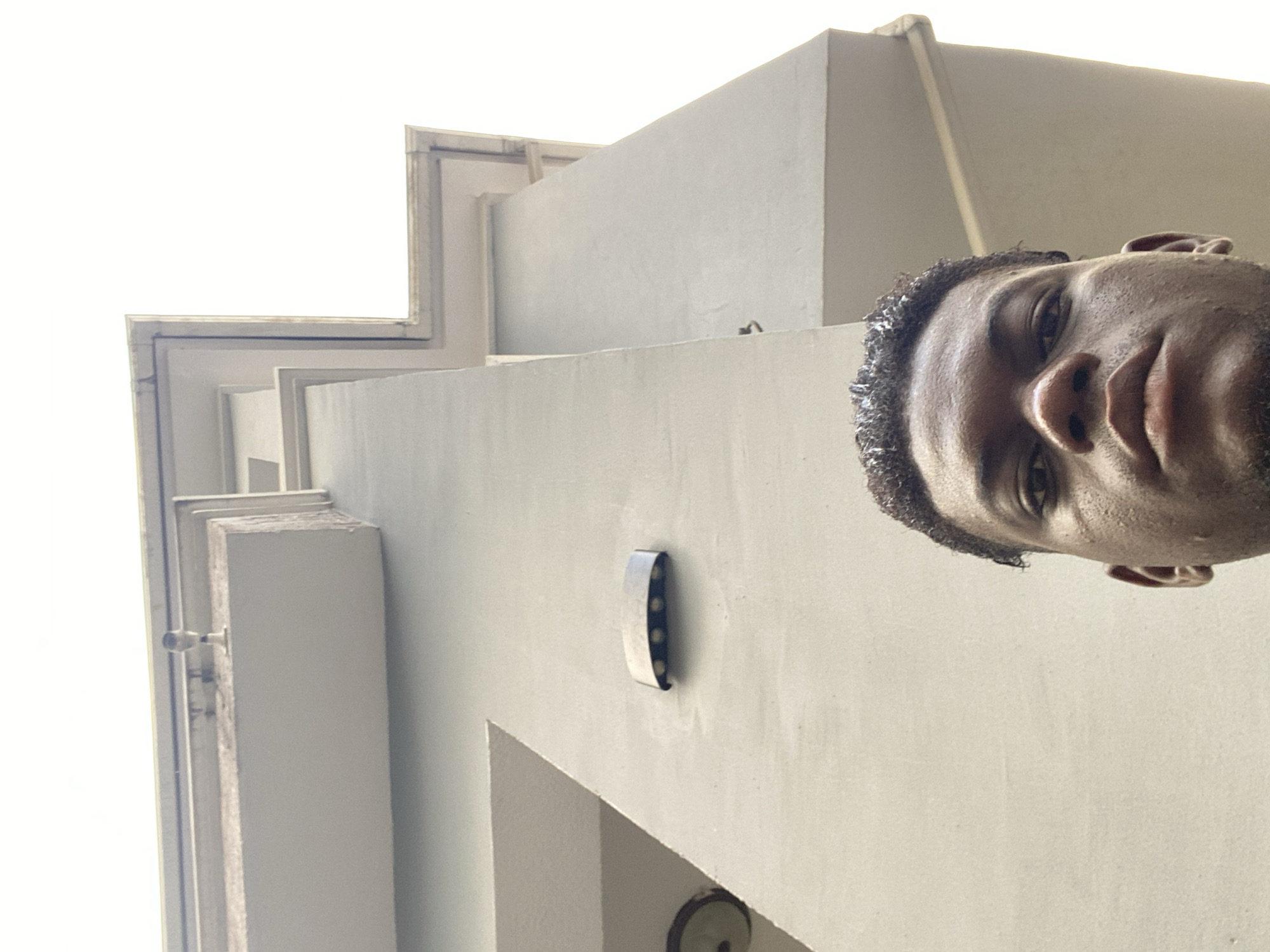 A low-angle shot shows a man's face looking down at the camera from a building exterior. The building is constructed of white concrete with sharp, angular lines. A light fixture and other small metallic fixtures are attached to the building's facade. The sky is visible in the upper-left corner, appearing bright and overcast.