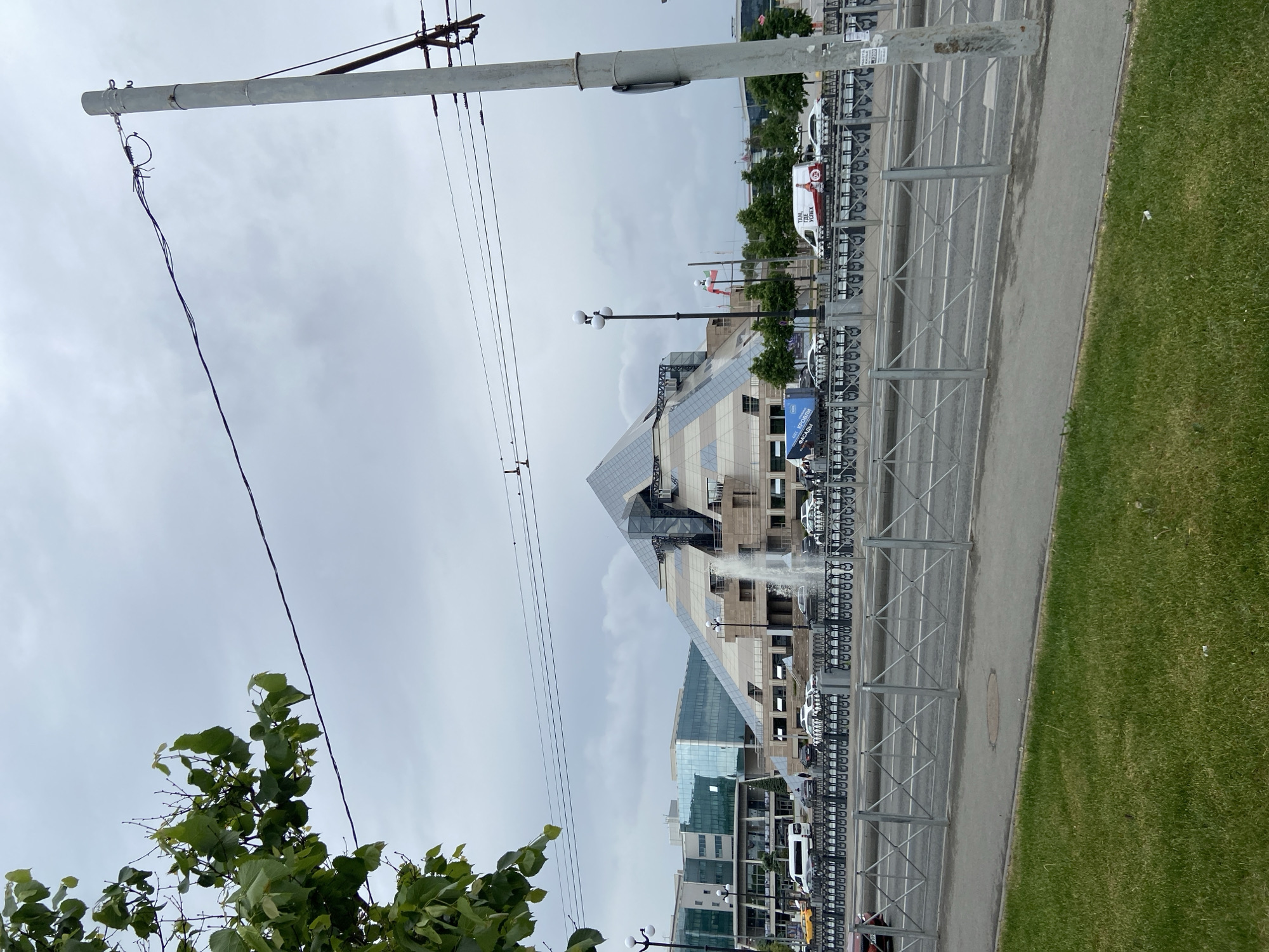The image captures a daytime urban scene in Kazan, Russia. The most prominent structure is a large building with a distinctive pyramid-like glass facade, possibly a public building or business center. A fountain with water spraying upwards is visible in front of the building. A row of parked vehicles lines the street adjacent to the building, with metal barriers separating them from the sidewalk and a grassy area to the right. Overhead, utility poles with power lines cross the frame. A portion of a tree with green foliage is visible in the lower left corner. The sky is overcast.