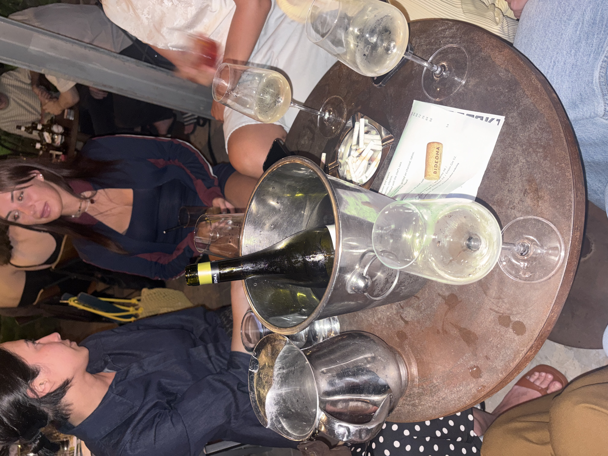 An outdoor seating area at a restaurant in Tel-Aviv, Israel. A group of people are gathered around a circular table, which is made of weathered metal. On the table, there are several glasses of white wine, an ice bucket with a bottle of white wine inside, a metal pitcher, an ashtray full of cigarette butts, and a menu. The people appear to be enjoying drinks and conversation. The overall atmosphere is casual and relaxed, suggesting a pleasant evening out.