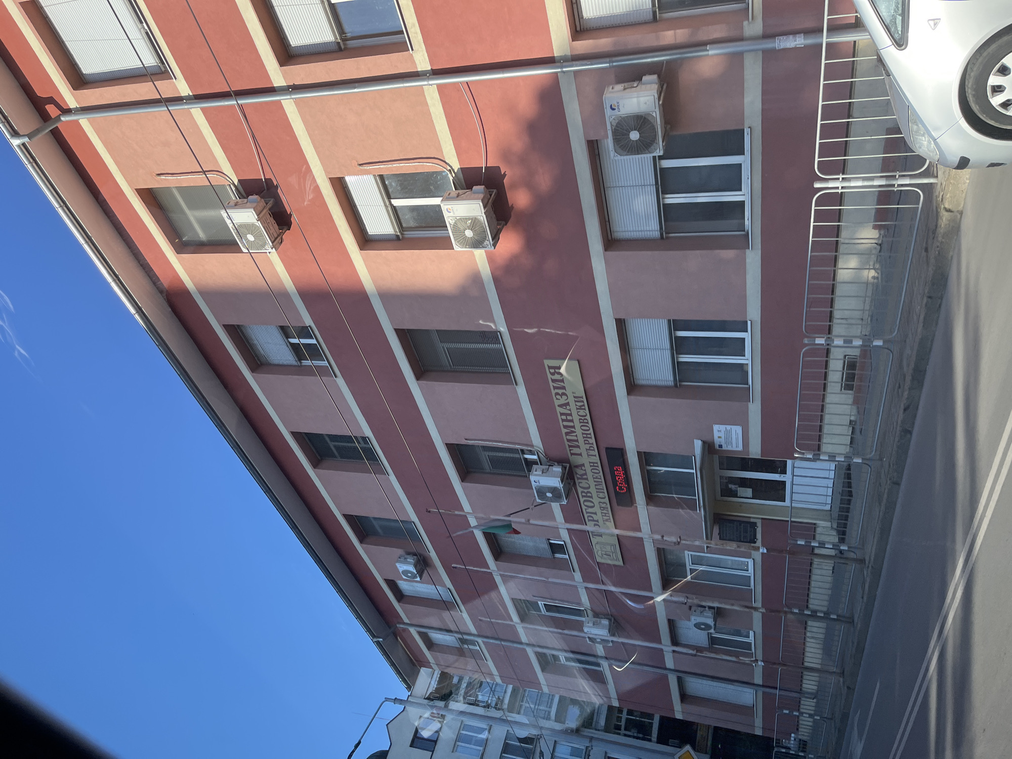The image shows the facade of a building, likely a school, in Stara Zagora, Bulgaria. The building is painted in pink and red horizontal stripes. There are multiple air conditioning units mounted on the exterior walls. Above the main entrance, there is a sign in Cyrillic script which reads "ТЪРГОВСКА ГИМНАЗИЯ" (Commercial High School) and below it, "КНЯЗ СИМЕОН ТЪРНОВСКИ" (Prince Simeon Tarnovski). There are also two Bulgarian flags on flagpoles. A digital display shows "Сряда" (Wednesday). The building is bordered by a metal fence and a street in the foreground. A silver car is partially visible on the right side of the image. The sky is clear and blue.