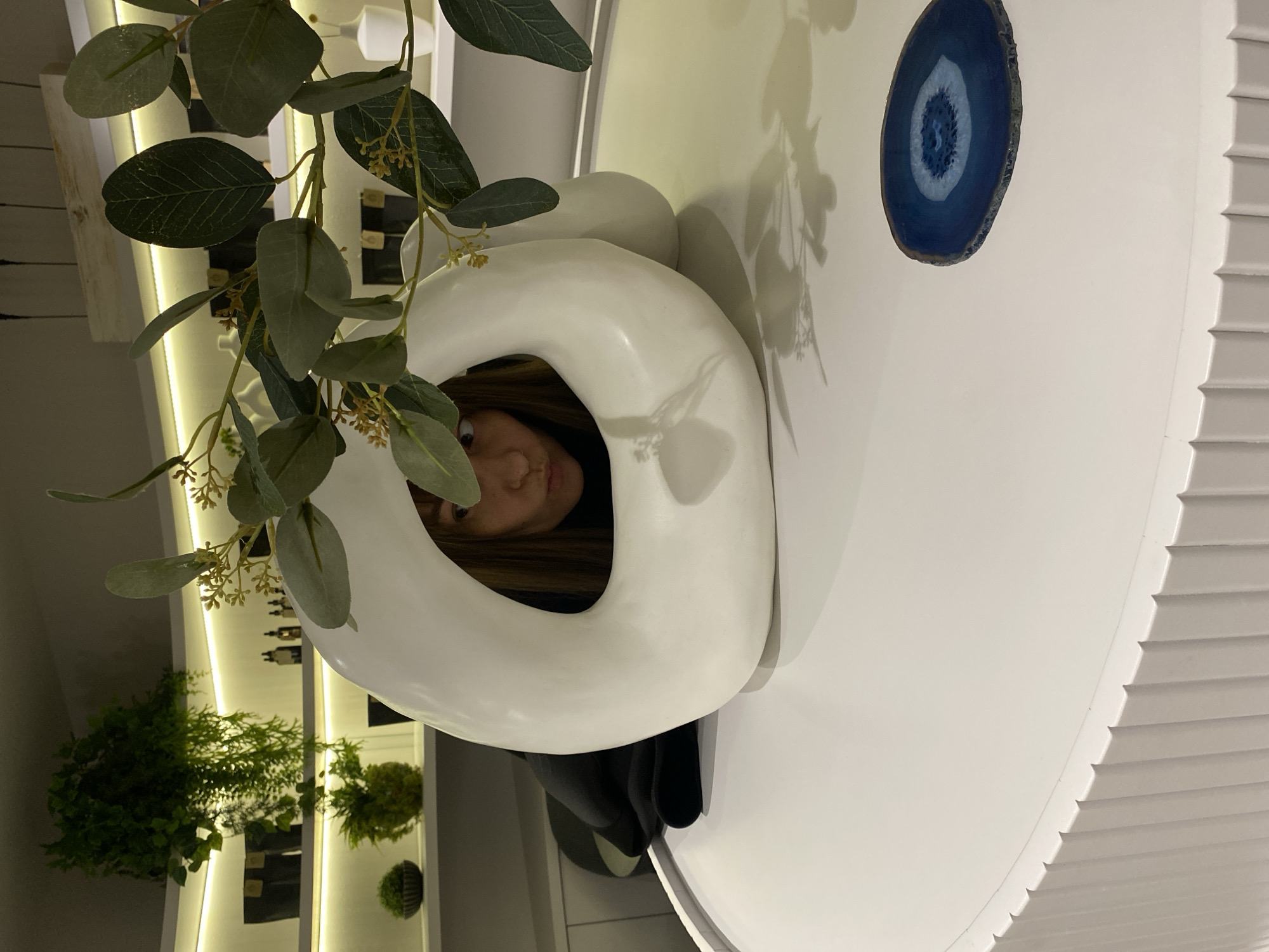 A person is peering through the center of a white, loop-shaped vase. The vase sits atop a white table with a blue and white stone disk nearby. A plant with green leaves is arranged around the vase. In the background, there's a shelving unit with bottles and small plants. The scene appears to be indoors. The location in Votkinsk, Russia, cannot be determined from the image.