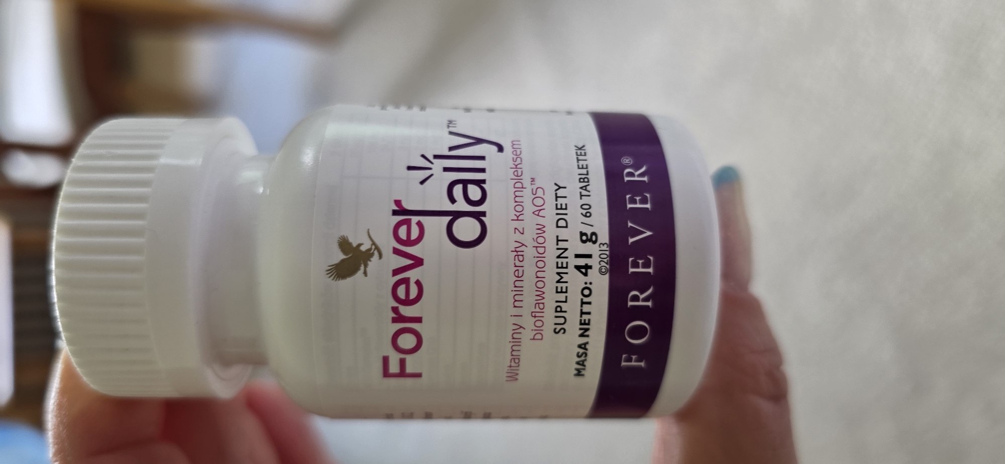 A white bottle of "Forever Daily" dietary supplements is held by a person's hand, with their thumb and forefinger obscuring the top of the bottle. The bottle has a white ribbed cap and a label with the brand name "Forever" in purple and "daily" in dark purple. Above the brand name is a silhouette of an eagle. The label also states "Witaminy i minerały z kompleksem bioflawonoidów AOS" (Vitamins and minerals with AOS bioflavonoid complex), "SUPLEMENT DIETY" (DIETARY SUPPLEMENT), "MASA NETTO: 41 g / 60 TABLETEK" (NET WEIGHT: 41 g / 60 TABLETS), and "©2013". A dark purple band at the bottom of the label features the word "FOREVER" in white text. The background is blurred and appears to be an indoor setting with wooden furniture. There is no information in the image to suggest a location in Maasbüll, Germany.