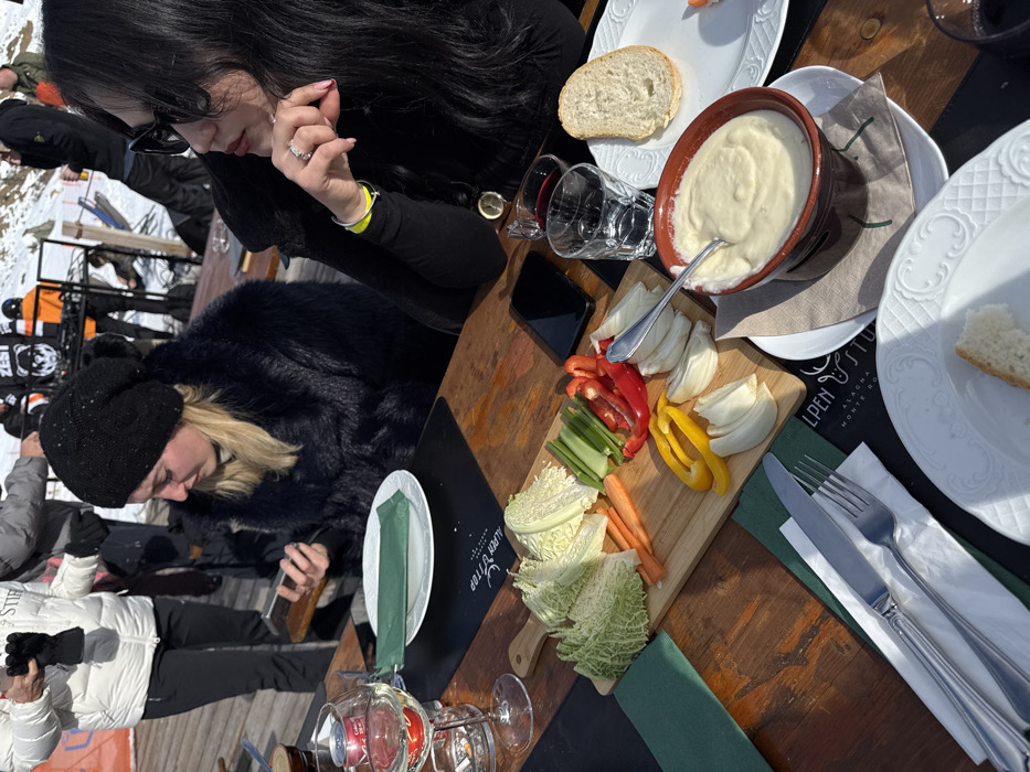 Two women are seated at a wooden outdoor table, enjoying a meal or drinks on a sunny day in a snowy mountain setting. The image is oriented vertically with the table angled diagonally across the frame.
The woman in the foreground on the left has blonde hair, wears a black beanie and a dark faux fur jacket, and is looking down at a smartphone in her hands. To her right, a woman with long dark hair and sunglasses is visible from the chest up. She wears a black top, has a silver ring on her left ring finger, and appears to be looking towards the table. Other individuals in winter clothing are partially visible in the snowy background, suggesting a ski resort environment.
On the table, a spread of food is laid out. A large brown bowl holds a creamy white dip, possibly fondue or a similar sauce, with a spoon resting in it. Beside it, slices of white bread are placed on a decorative white plate. A wooden serving board features a colorful array of raw, sliced vegetables including red and yellow bell peppers, celery sticks, carrot batons, sliced fennel, and two piles of chopped savoy cabbage or lettuce.
Two wine glasses are visible, one containing a red liquid and another clear glass. White plates, silverware (a fork and knife), and green napkins are arranged on the table. A black smartphone is also resting on the table near the dark-haired woman.
Visible text on a black placemat or menu item reads "ALPEN TOP" with a logo resembling a deer or ibex head, followed by "ALAGNA" and partially "MONTE RO**", confirming the location as Alagna Valsesia, Italy, as provided in the context. The bright daylight casts distinct shadows, indicating a clear, sunny sky, likely midday or early afternoon. The overall atmosphere appears relaxed and social in a winter mountain resort.