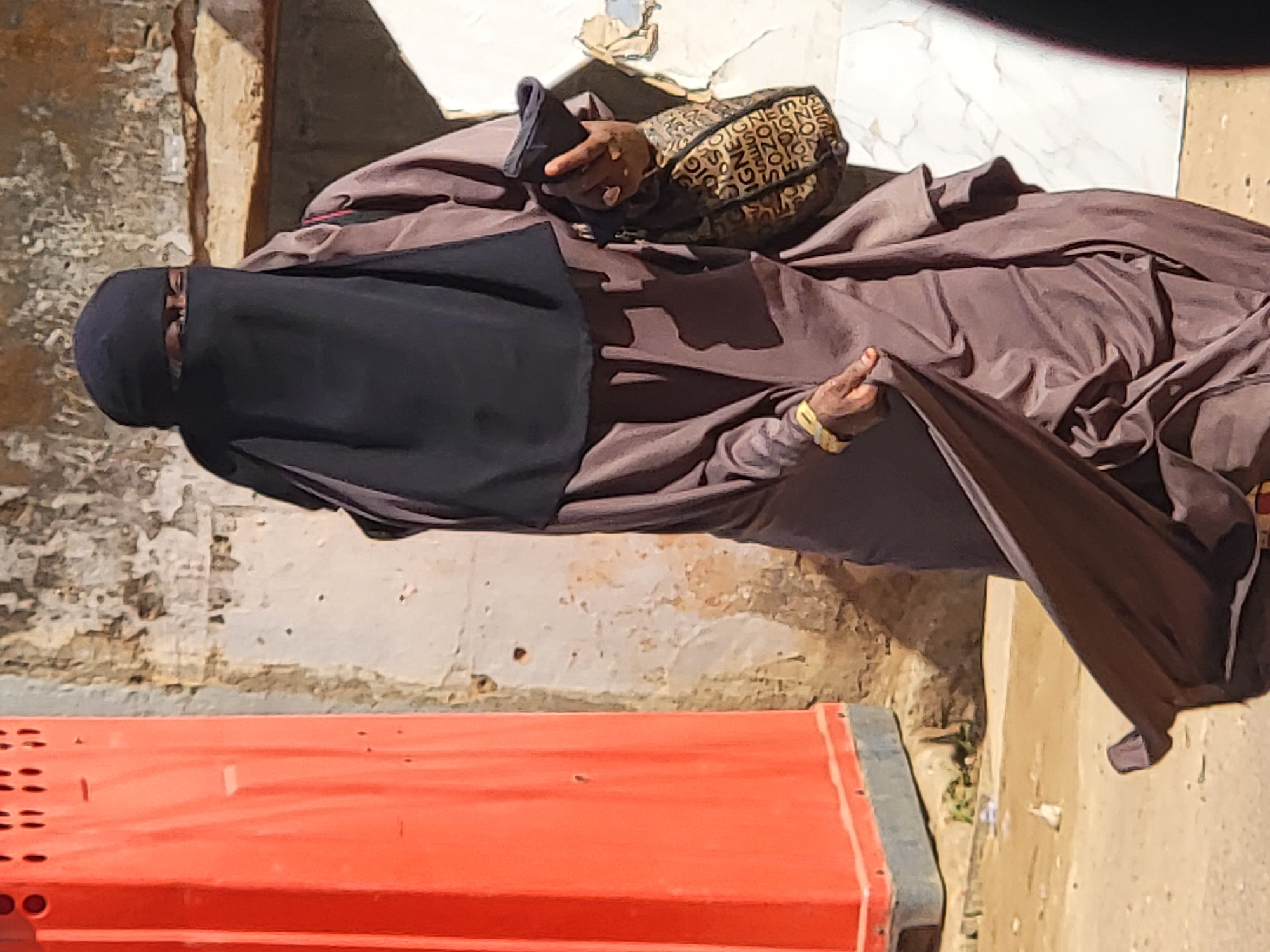 The image captures a person, likely female, standing outdoors in Maiburiji, Nigeria. The individual is dressed in a loose-fitting, full-length brown abaya and a black niqab that covers the face, leaving only the eyes visible. Dark skin is observable on the hands and around the eyes.
On the person's right arm, a patterned sleeve or bag is seen, featuring repeating text fragments such as "SIGN," "GNE," and "SOZO." The right hand holds a small, dark object, possibly a mobile phone or a wallet. A yellow or gold-colored bracelet adorns the left wrist.
The background consists of a textured wall with light brown and beige tones, showing signs of wear with peeling paint or plaster. The ground is a light, dusty color, possibly concrete or packed earth. In the lower foreground, a large, bright orange object, possibly a container, barrier, or part of a piece of equipment, is partially visible on the left. It has visible perforations and straight lines.
The scene is brightly illuminated by natural sunlight, casting distinct shadows, indicating a midday or early afternoon setting with clear weather. The person's posture appears casual, and the visible eye area suggests a calm demeanor. The overall impression is an everyday street or outdoor scene within an urban or semi-urban context.